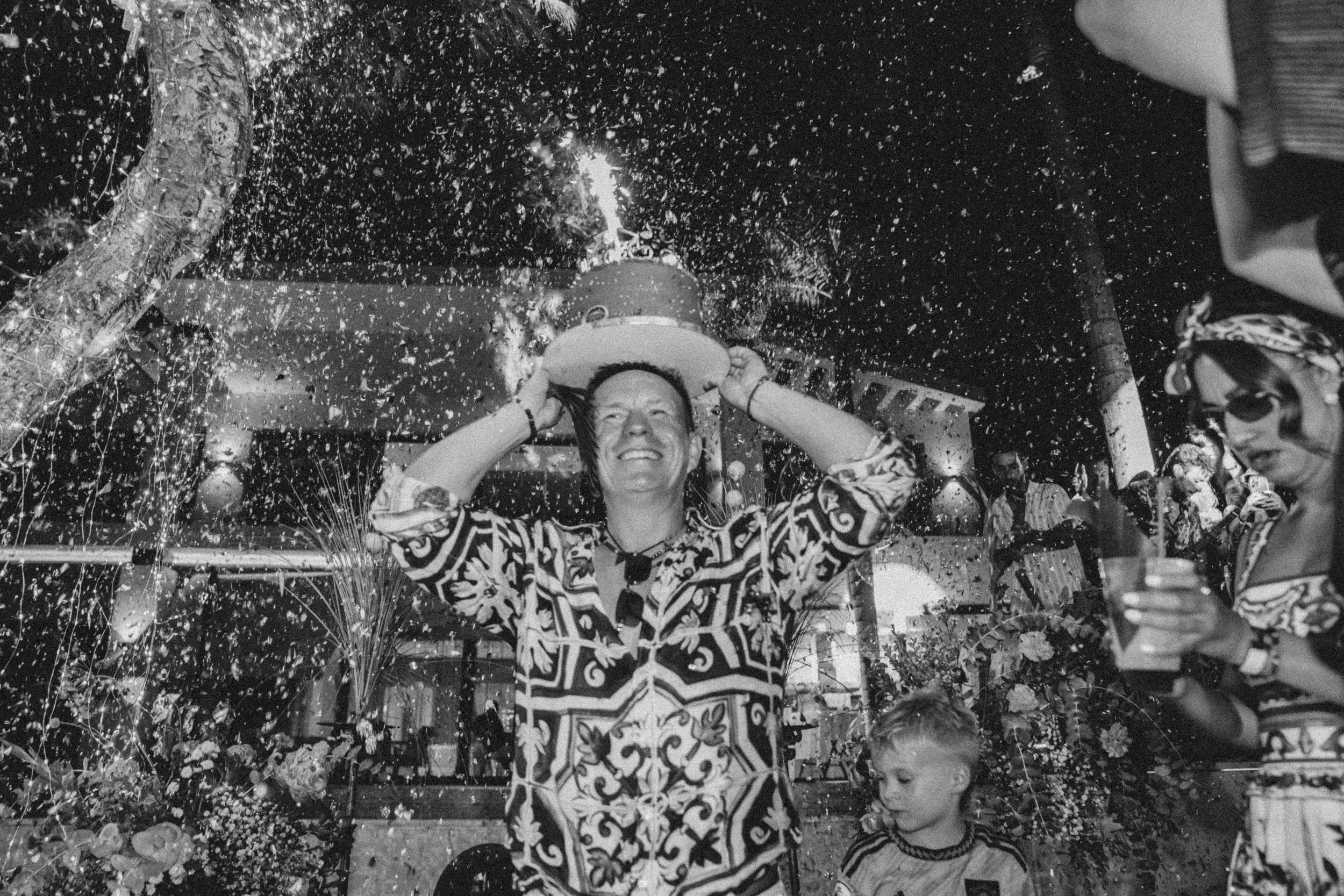 A woman smiling and holding a hat above her head, surrounded by confetti and people at a celebration or party.