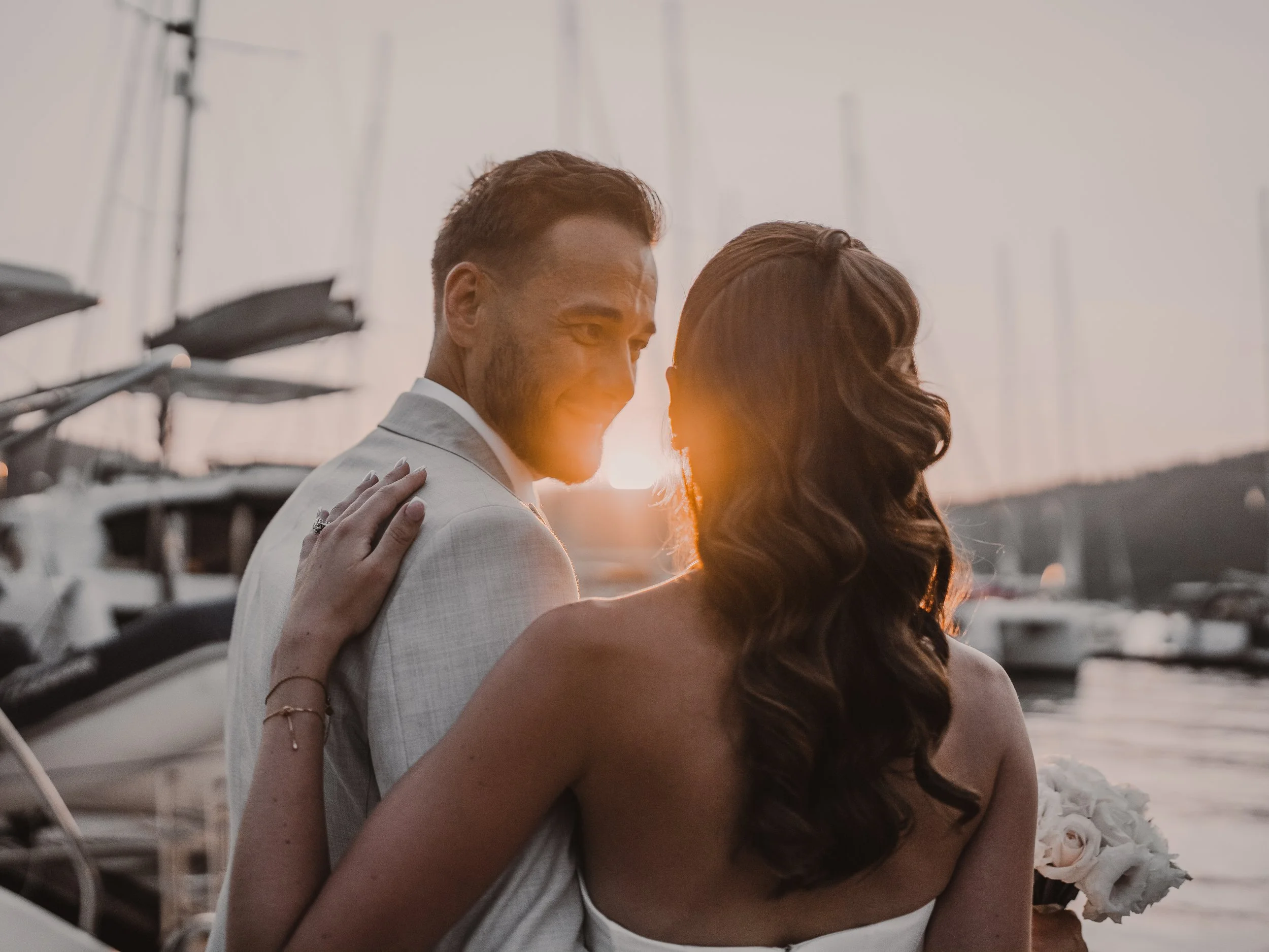 A couple dressed in formal attire embracing at sunset on a beach, with sailboats in the background.