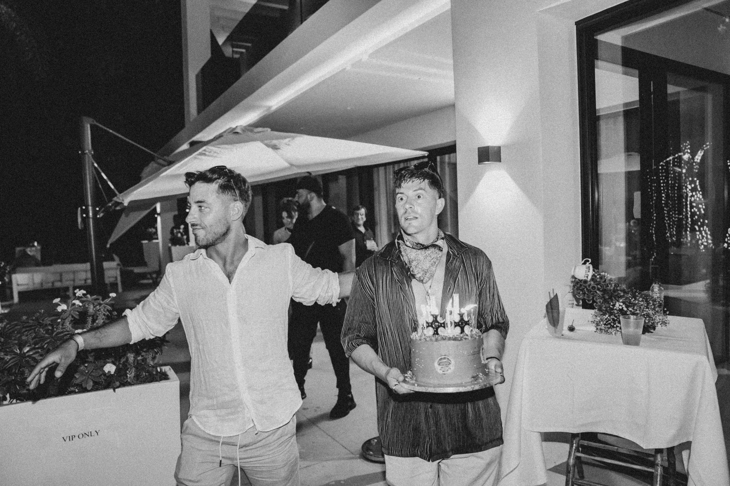 Two men are at a birthday celebration, with one holding a cake with candles, in an indoor setting with some people and decorations around.
