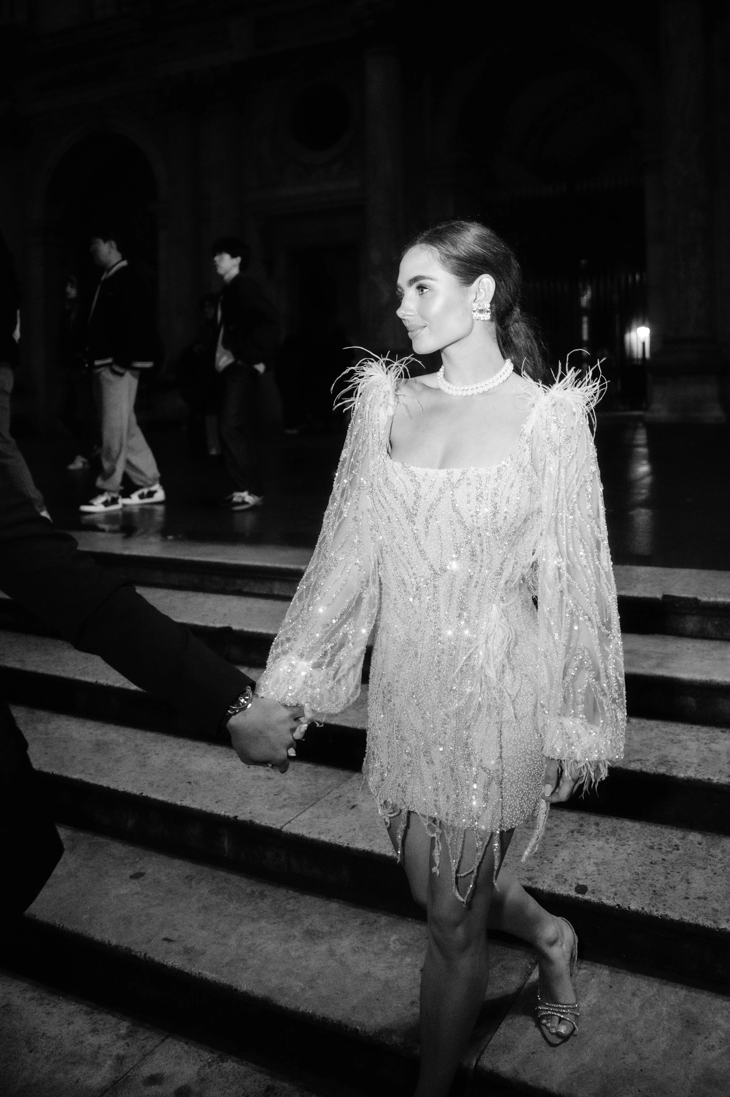 A woman in a glamorous, beaded dress holding hands with someone, walking down the stairs at an event. She wears jewelry and has dark hair styled back.