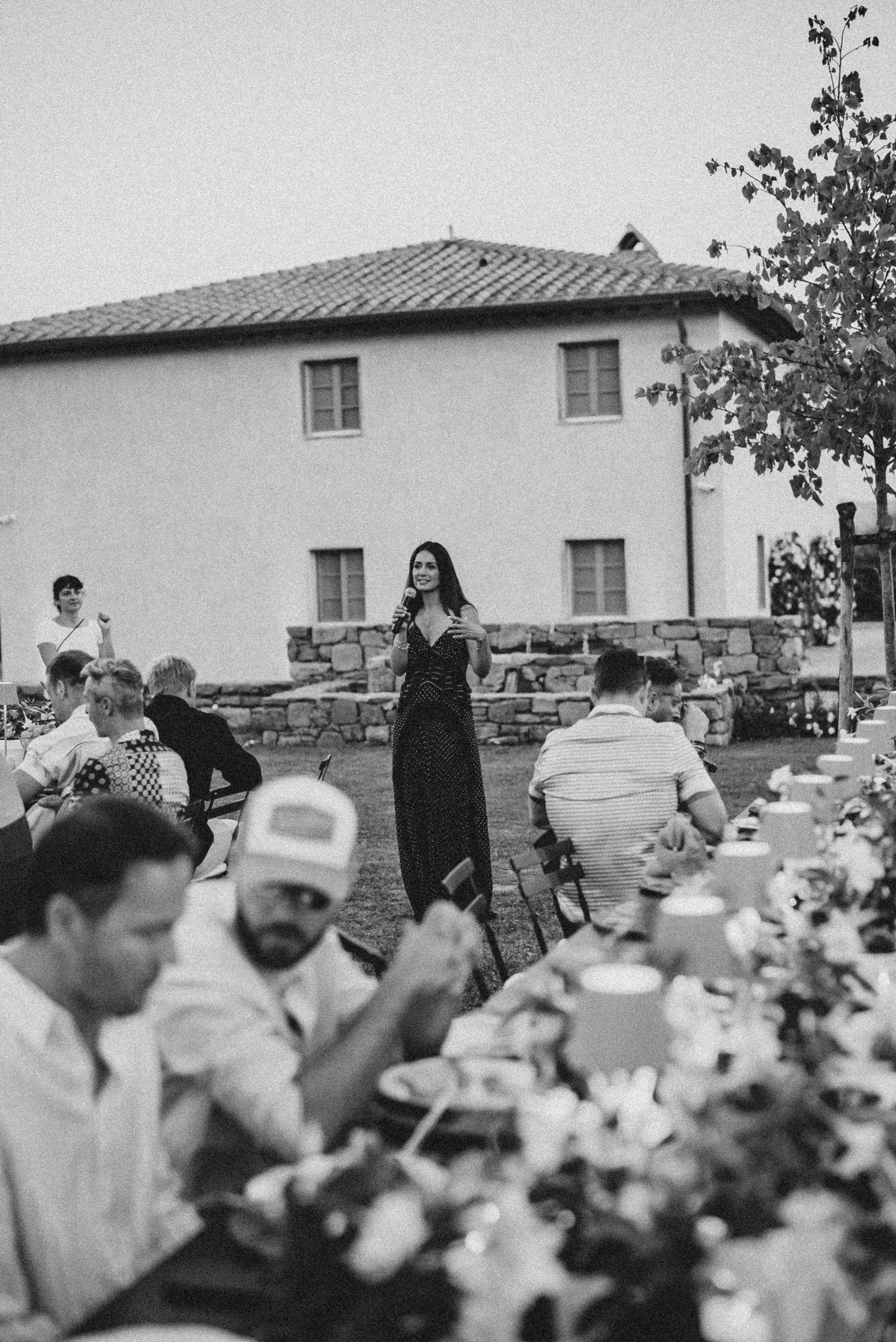 A woman holding a microphone and speaking at an outdoor gathering, with people seated at tables, in front of a building with a tiled roof and trees.