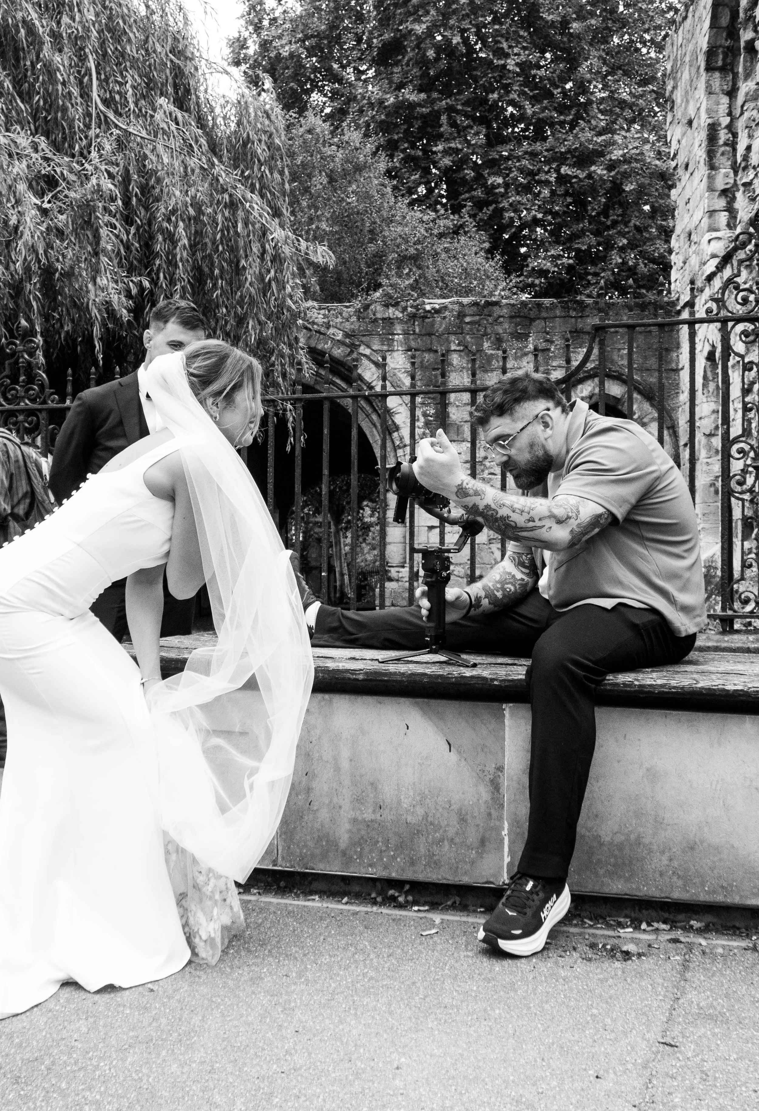 Black and white photo of a wedding photographer taking pictures of a bride in her wedding dress. They are outdoors, with a brick wall, iron fence, and trees in the background.