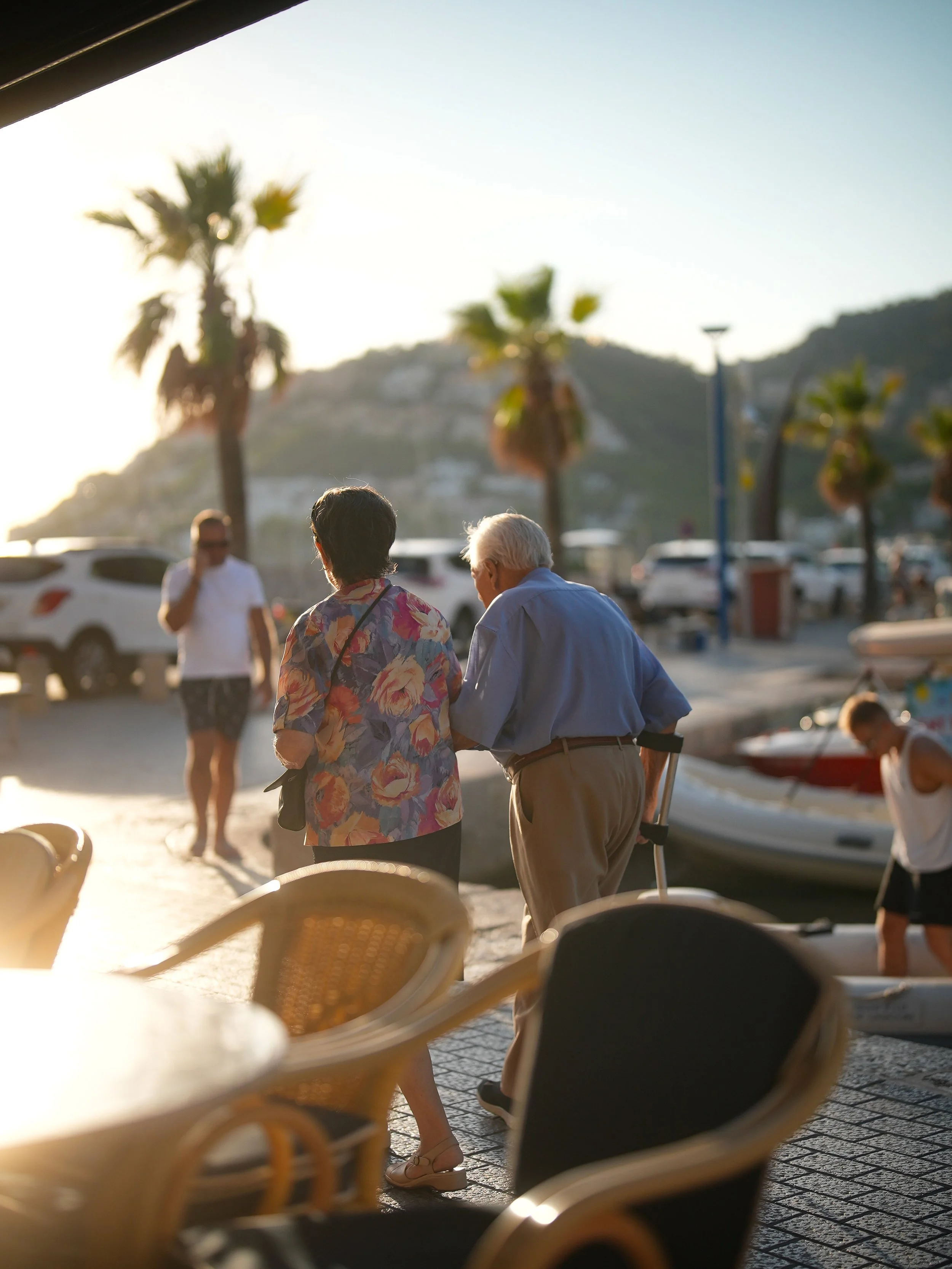 People walking along a waterfront promenade with palm trees and boats in the background, viewed from behind a row of empty chairs.