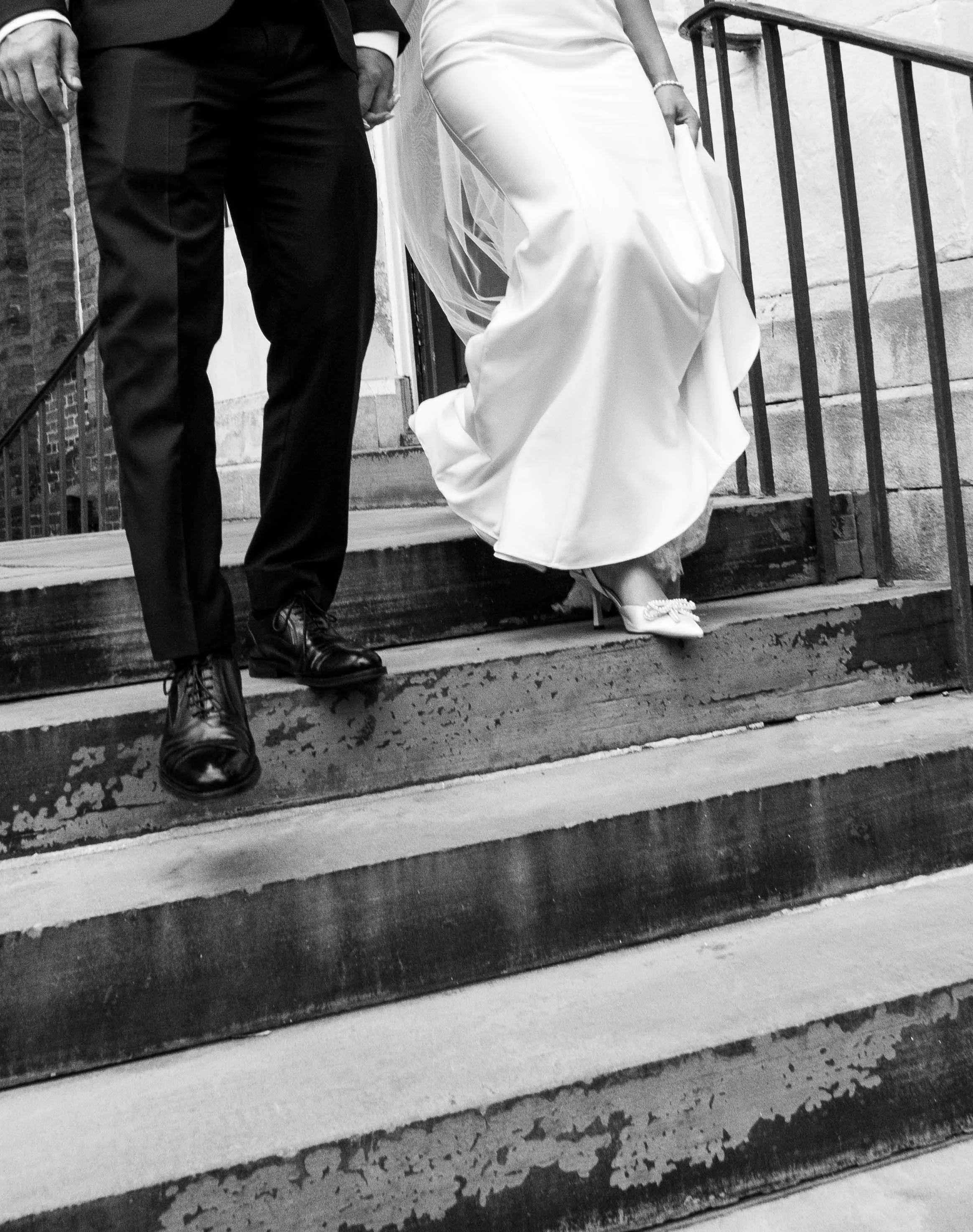 Close-up black and white photo of a man and a woman standing on stairs, dressed in formal attire, with the woman's dress and shoes visible.