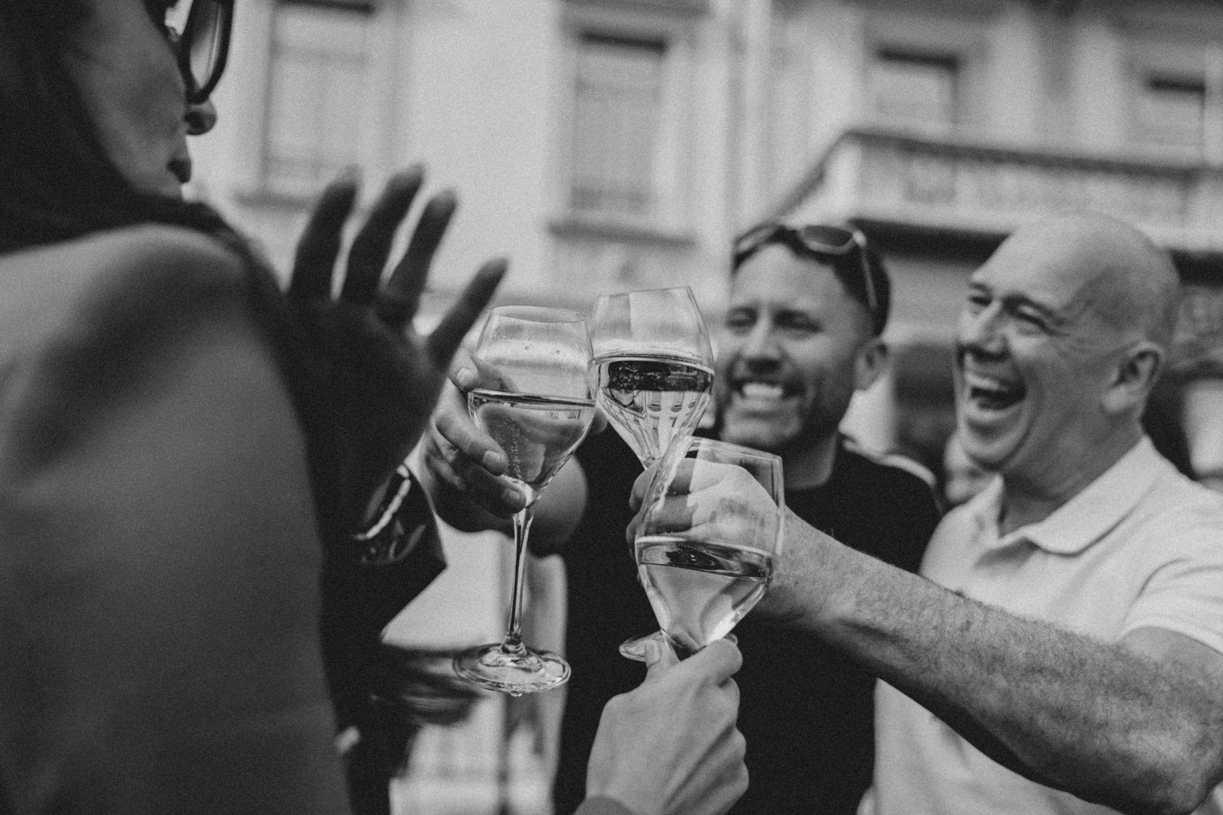 Four people clinking wine glasses in a toast, smiling, gathered outdoors with buildings in the background.