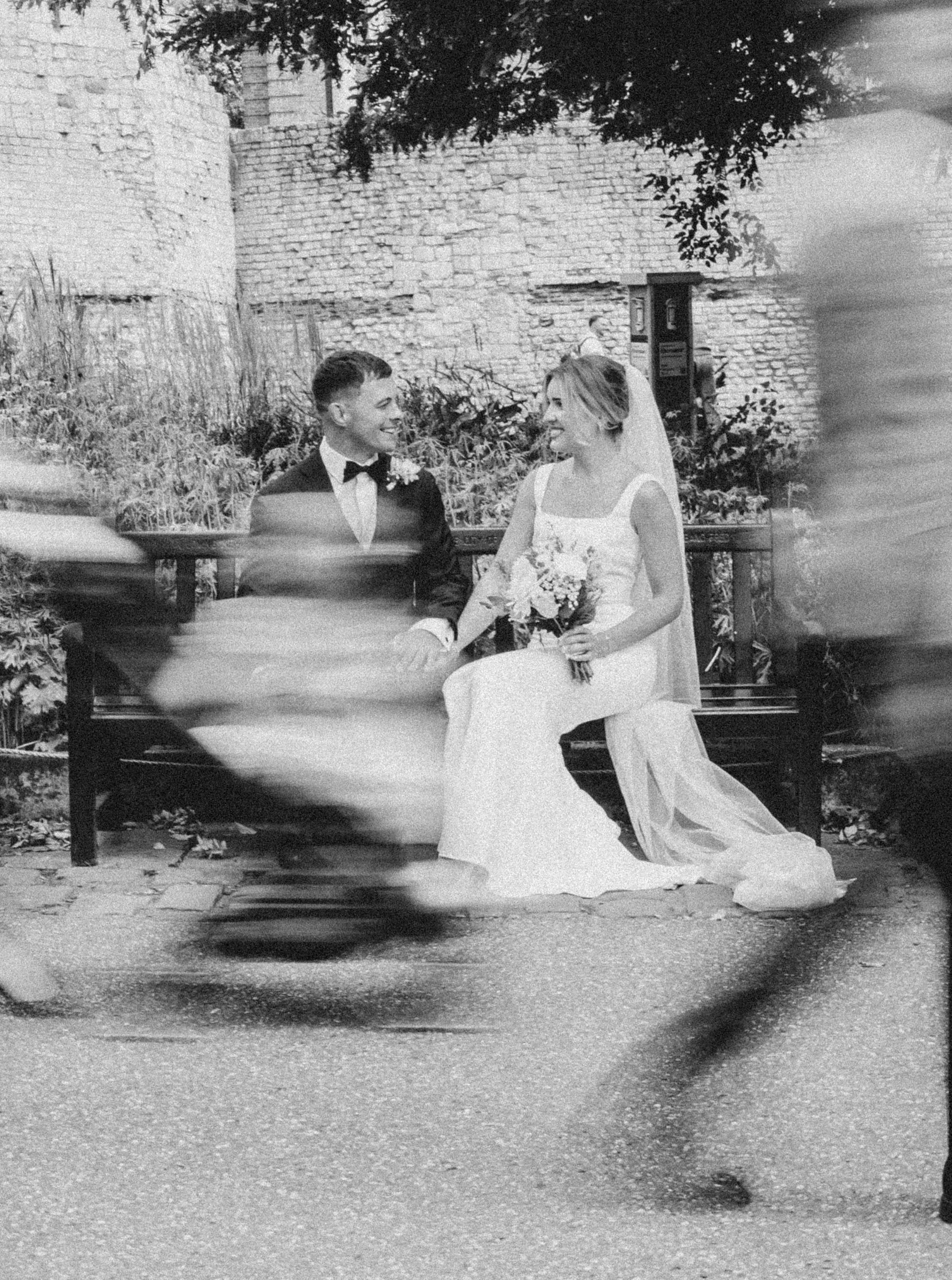 A black and white photograph of a bride and groom sitting on a park bench, smiling at each other, with a brick wall and trees in the background.