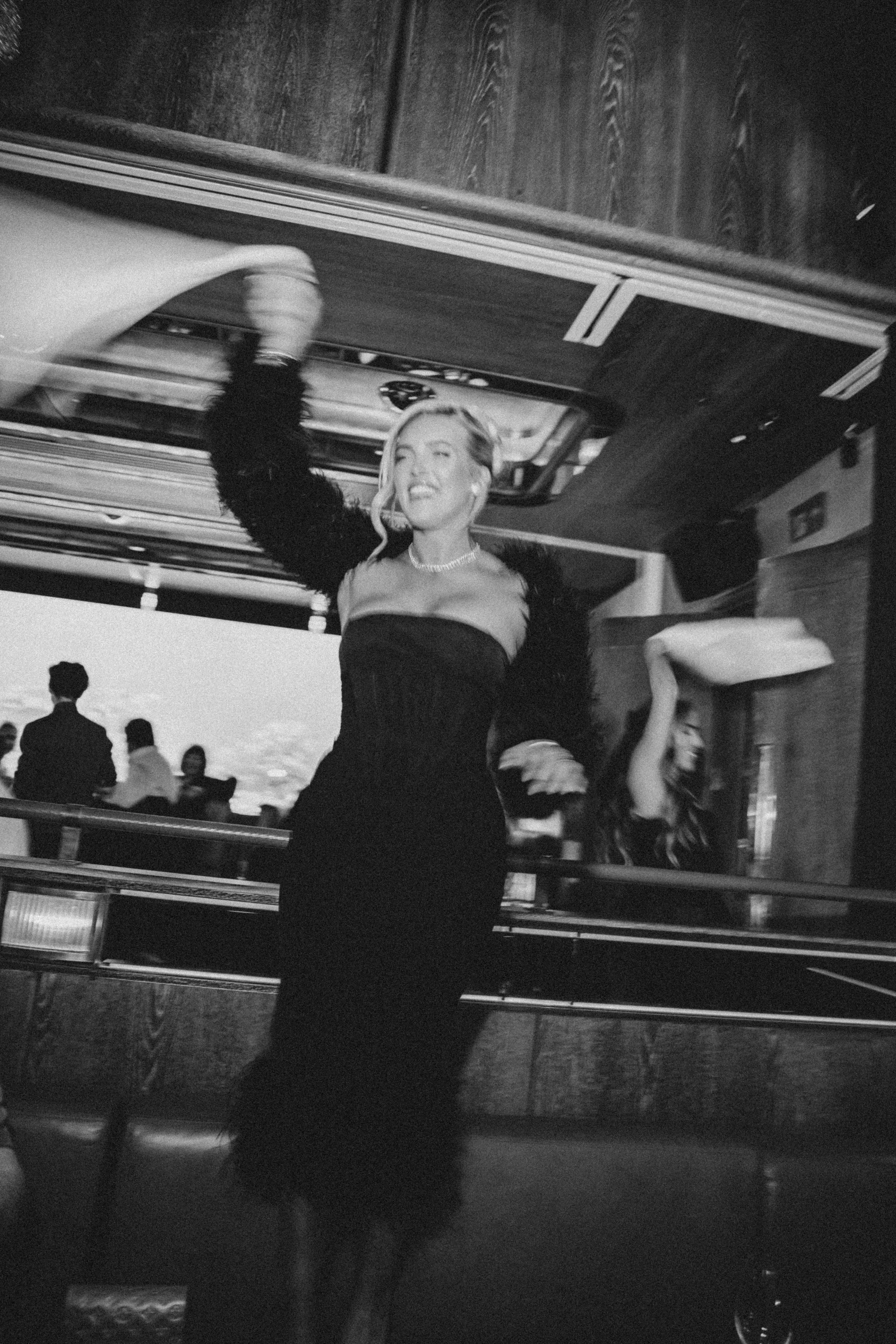 A woman with long hair wearing a black dress and necklace, smiling and throwing her arm up at an indoor event or party. There are other people in the background, some sitting and some standing.