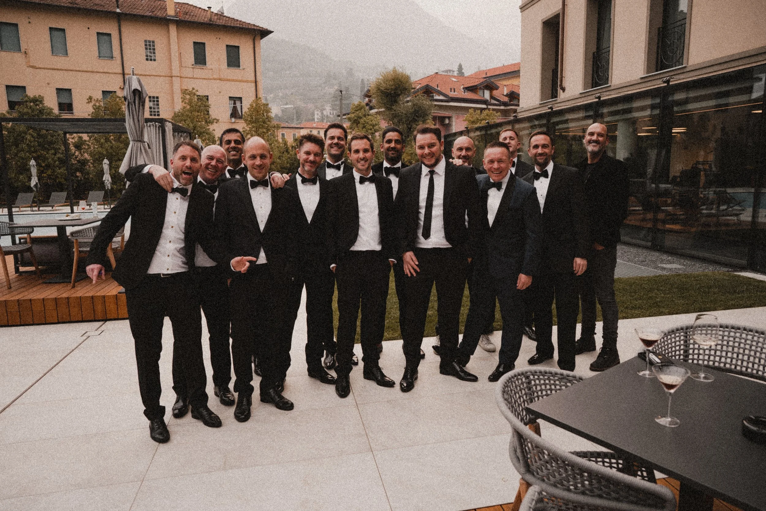 A group of men dressed in tuxedos and bow ties standing together outdoors, smiling for a photo during a celebration or formal event, with buildings and trees in the background.