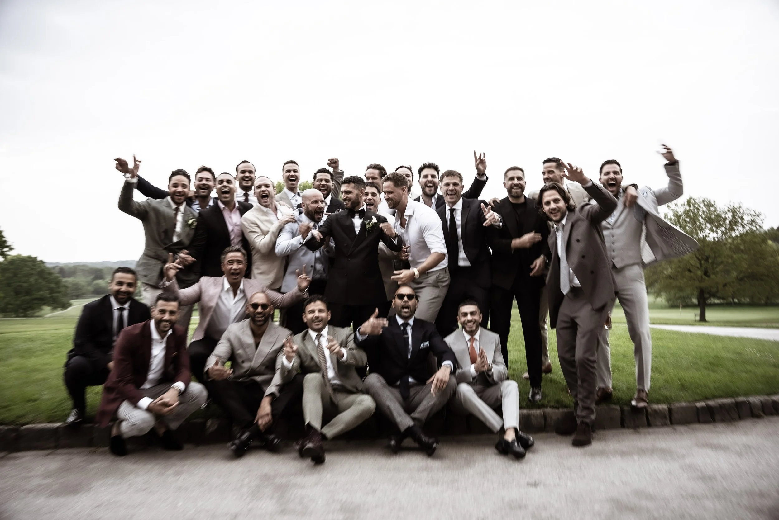 A large group of men dressed in suits posing outdoors, with most smiling, some making gestures, and a cloudy sky in the background.