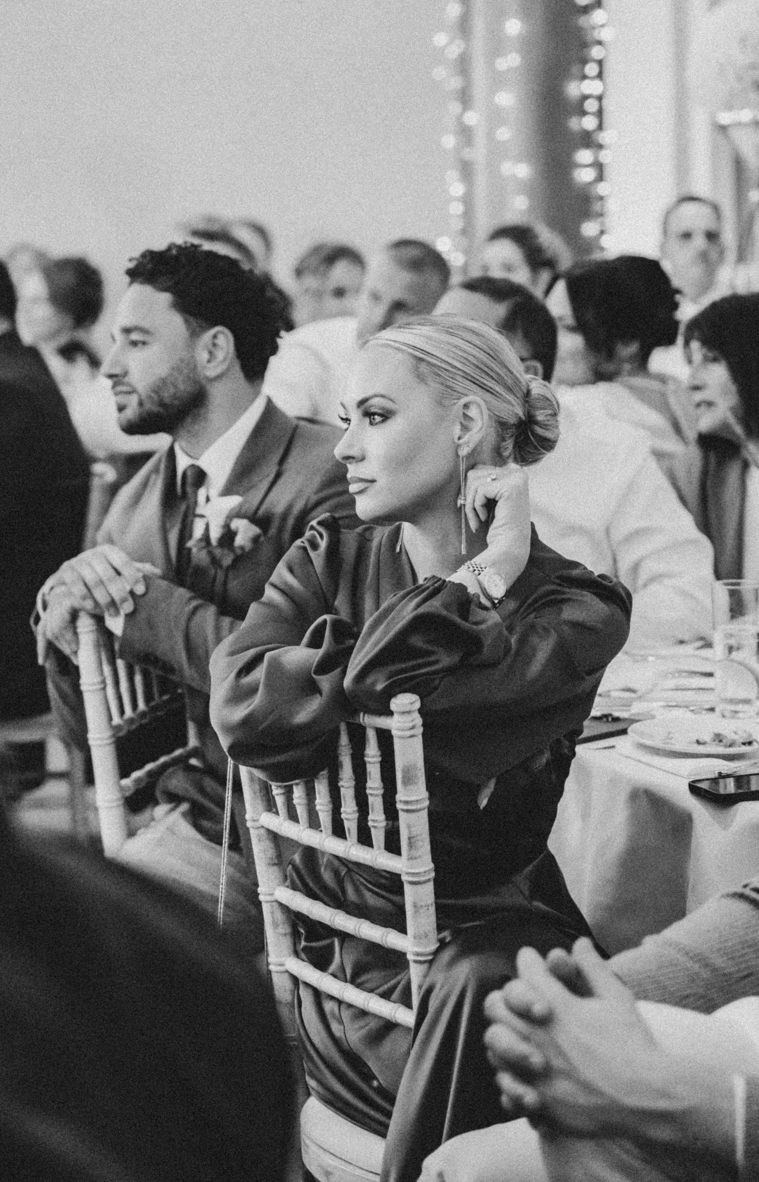 Black and white photo of people attending a formal event, seated at a banquet table, with a woman with blonde hair and a man with dark hair in the foreground. The woman has her hand resting near her face and is wearing elegant attire and jewelry. The