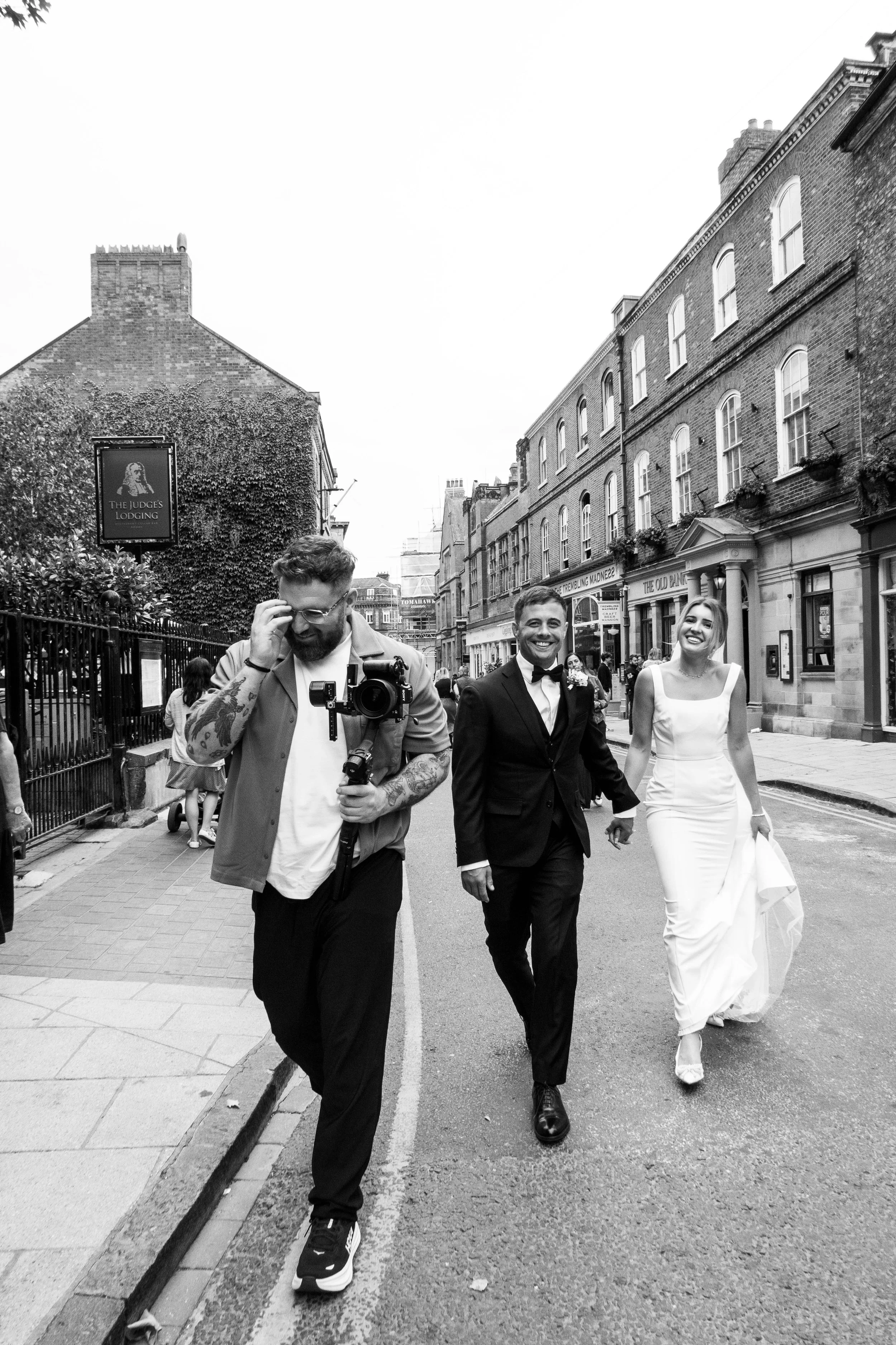 A black and white photograph of a couple in wedding attire walking down a city street, accompanied by a man taking a photo of them. The groom is in a tuxedo, and the bride is in a white wedding dress. They are smiling. The street features multi-story
