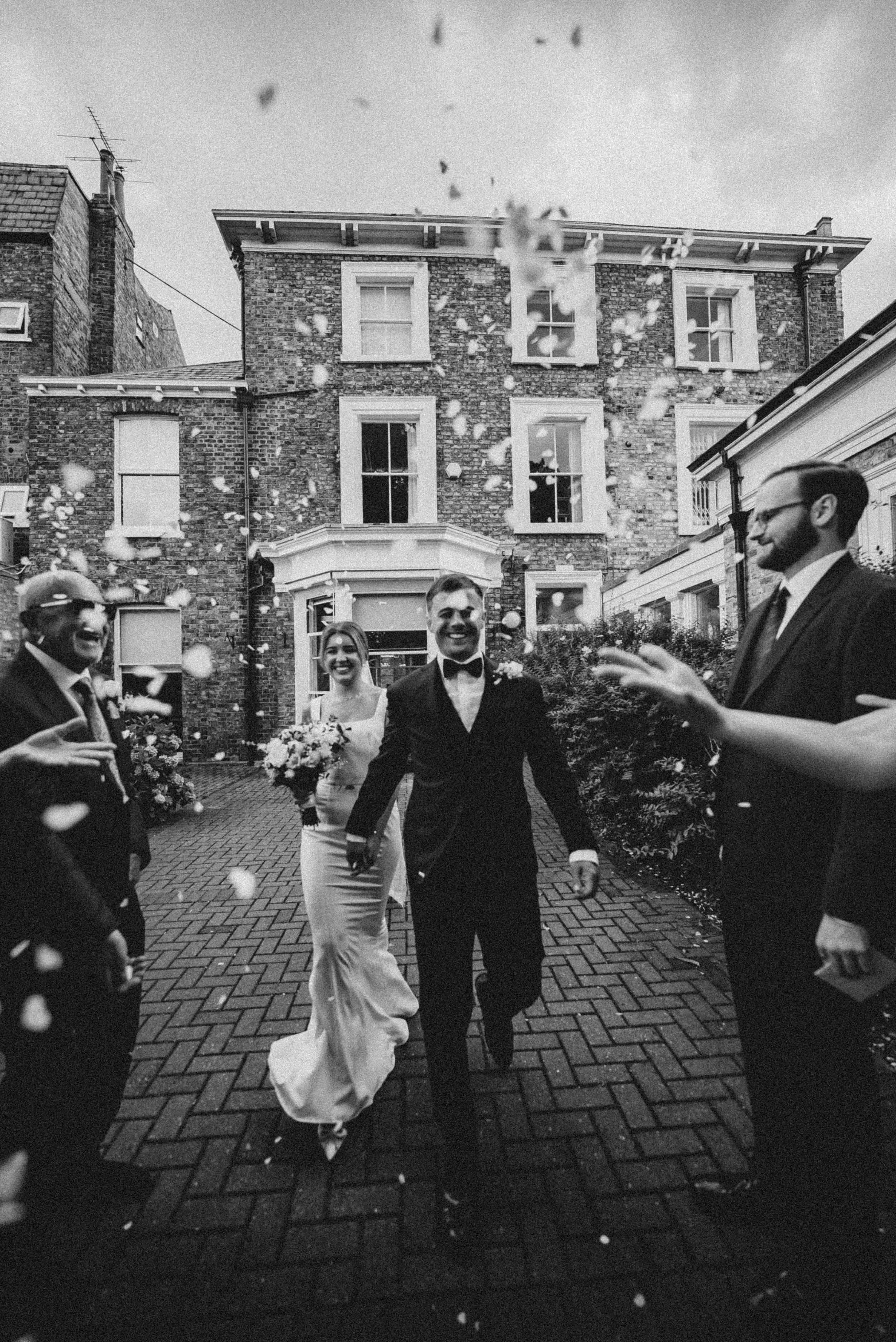 Black and white photograph of a wedding celebration with a bride and groom walking together outside a brick building, surrounded by guests throwing flower petals, celebrating.