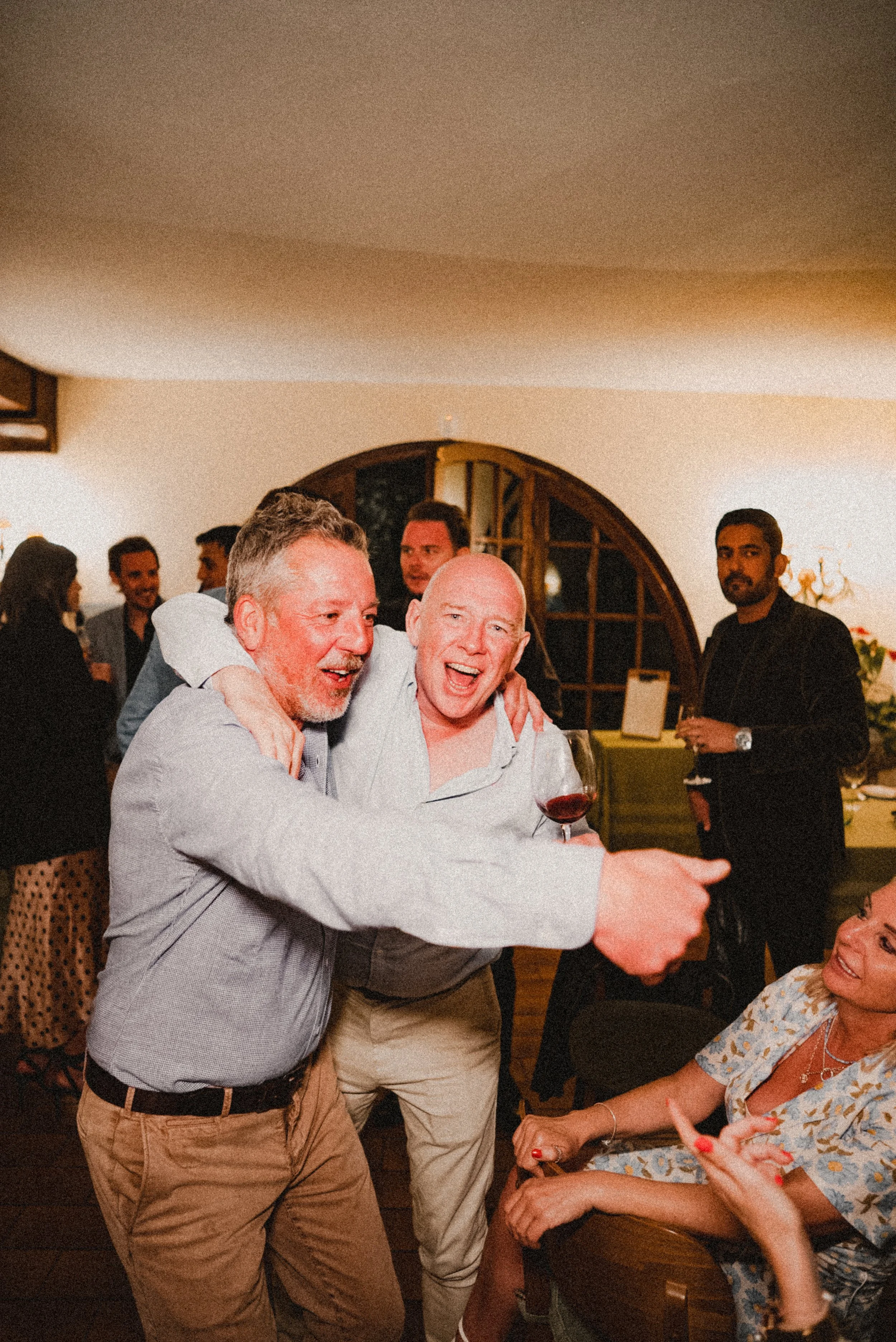 A group of people at a social gathering smiling, with two men in the foreground embracing and holding wine glasses, and others standing in the background.
