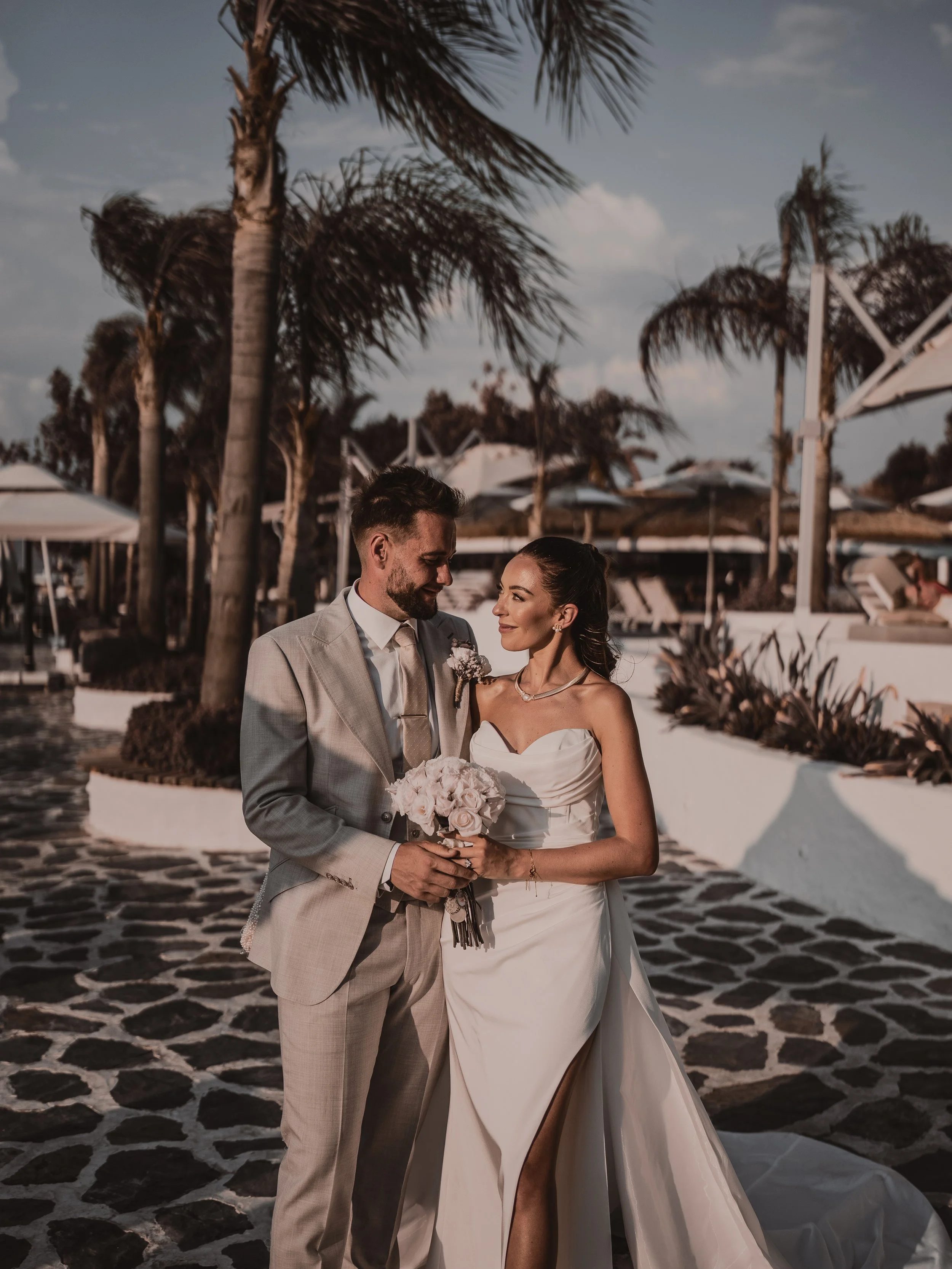 A bride and groom standing close together outdoors at sunset, with palm trees and lounge chairs in the background, smiling and looking at each other, dressed in wedding attire.
