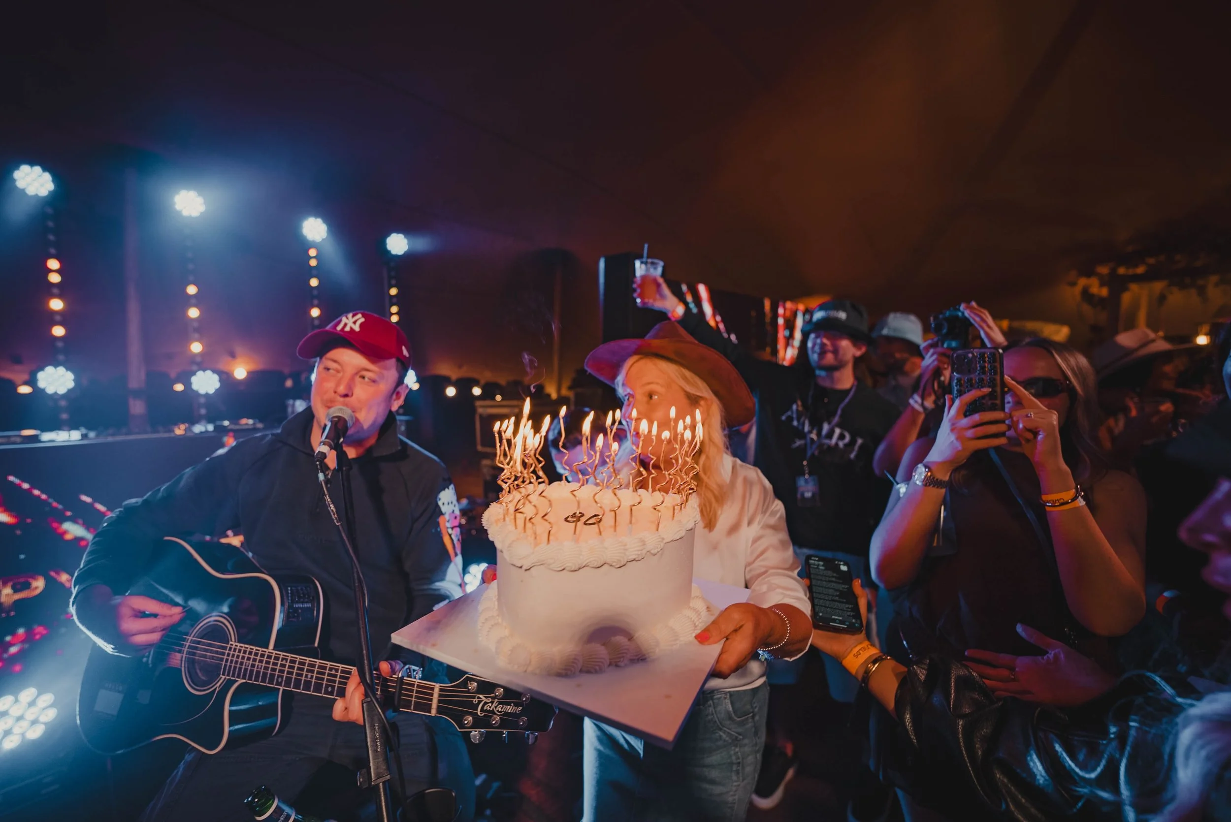 A woman with blonde hair wearing a large hat and a white sweater holding a birthday cake with lit candles. A man with a red baseball cap playing an acoustic guitar and singing into a microphone. Several people in the background, some taking photos an