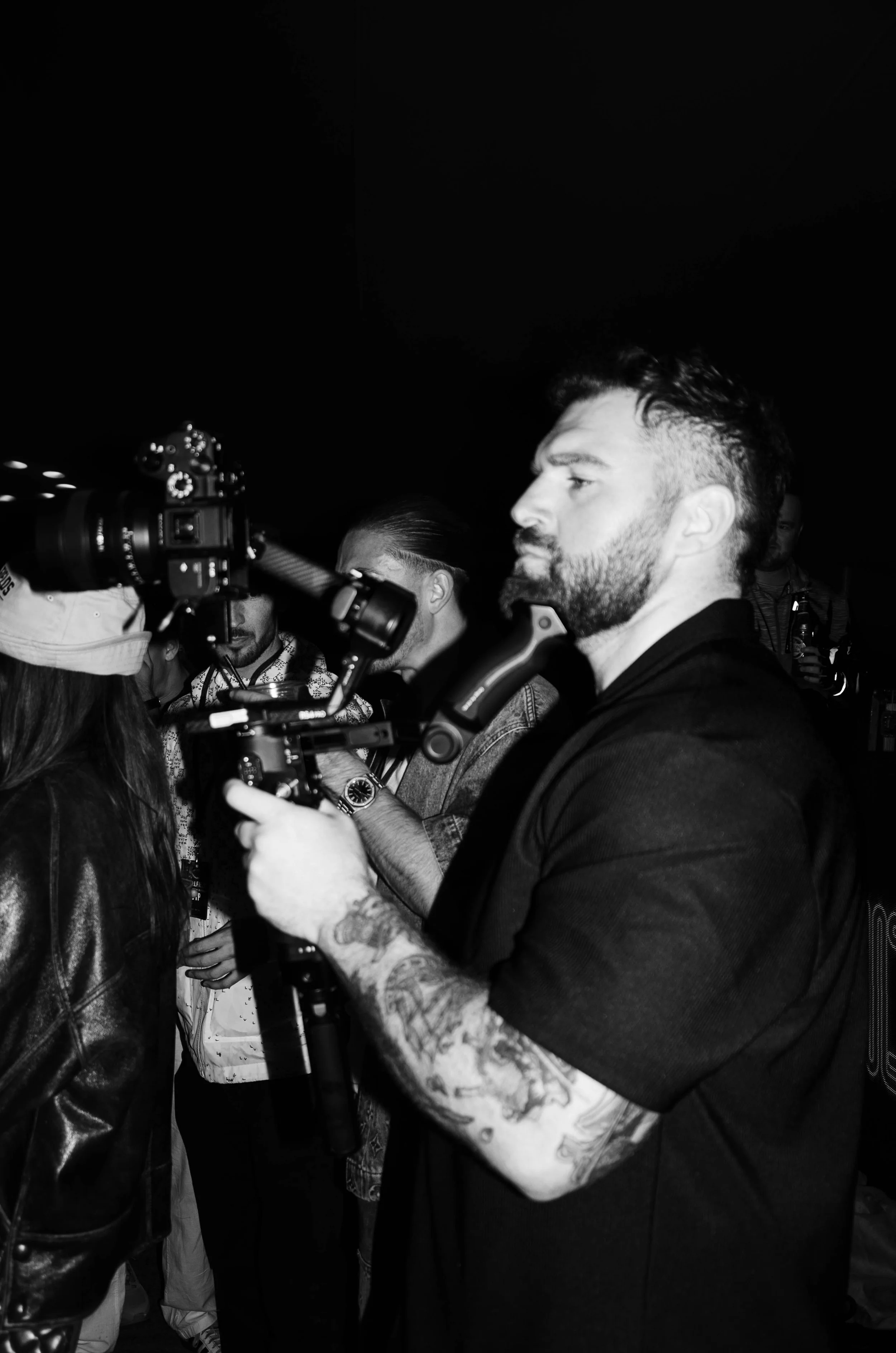 A man with a beard and tattoos on his arm is holding and operating a camera. Several other people are gathered around, some looking at the camera, in a dark indoor setting.