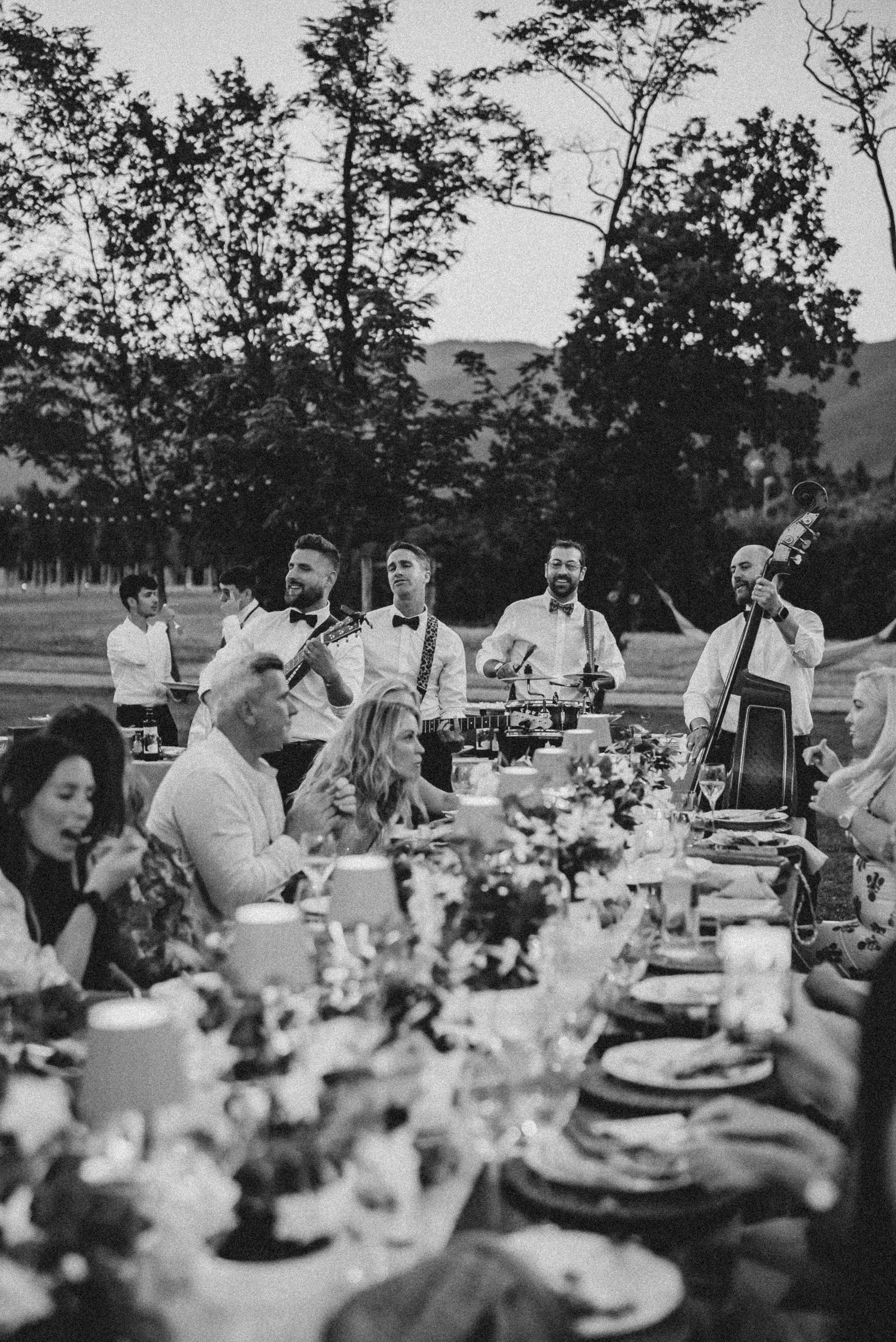 A black and white photo of a group of musicians performing at an outdoor event, with tables of people dining in the foreground and trees in the background.