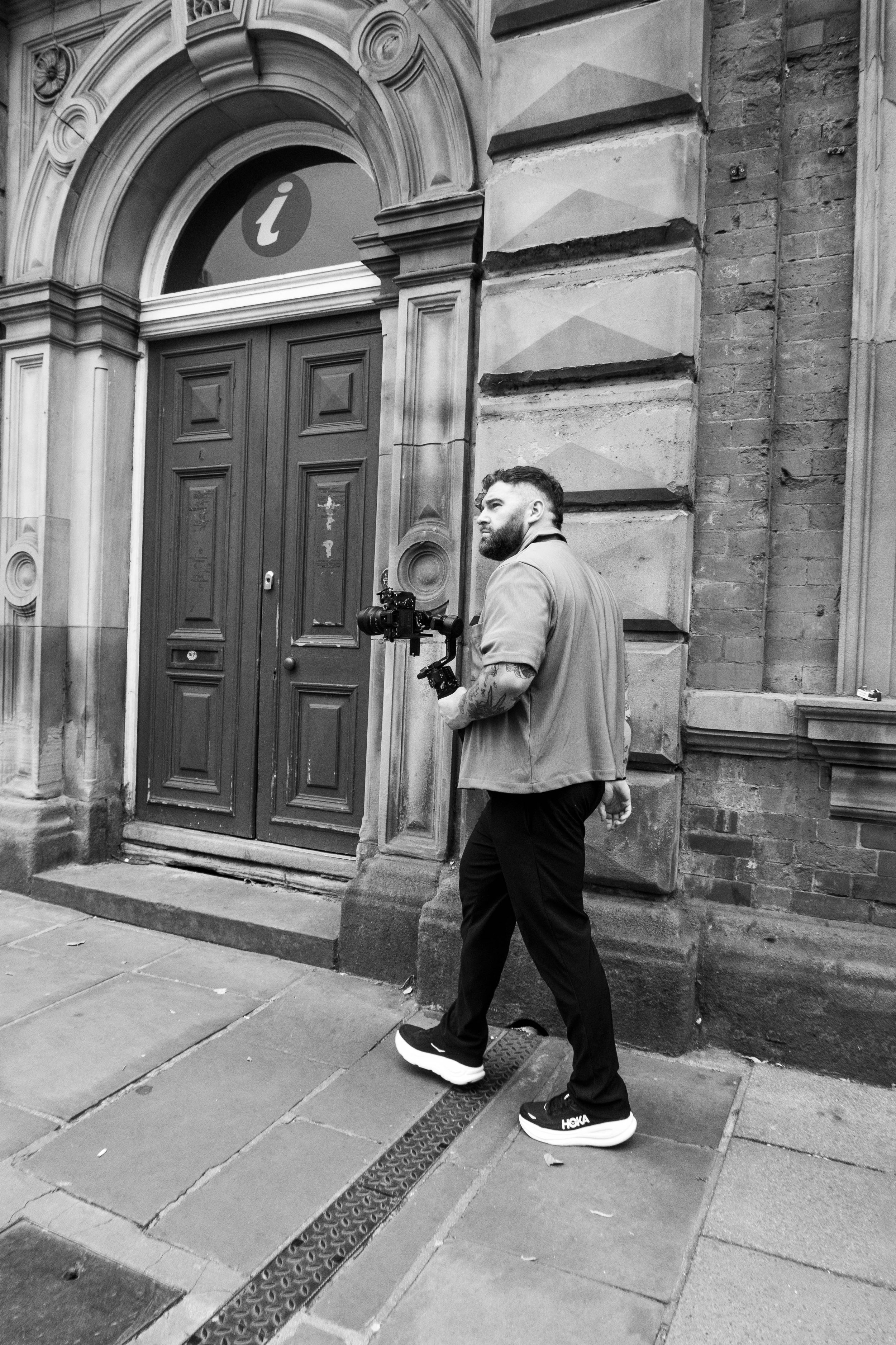 Black and white photo of a man with a beard walking past a building with arched front door and ornate stone facade. The man is wearing a jacket, dark pants, and Hoka sneakers, and is carrying a camera in one hand.