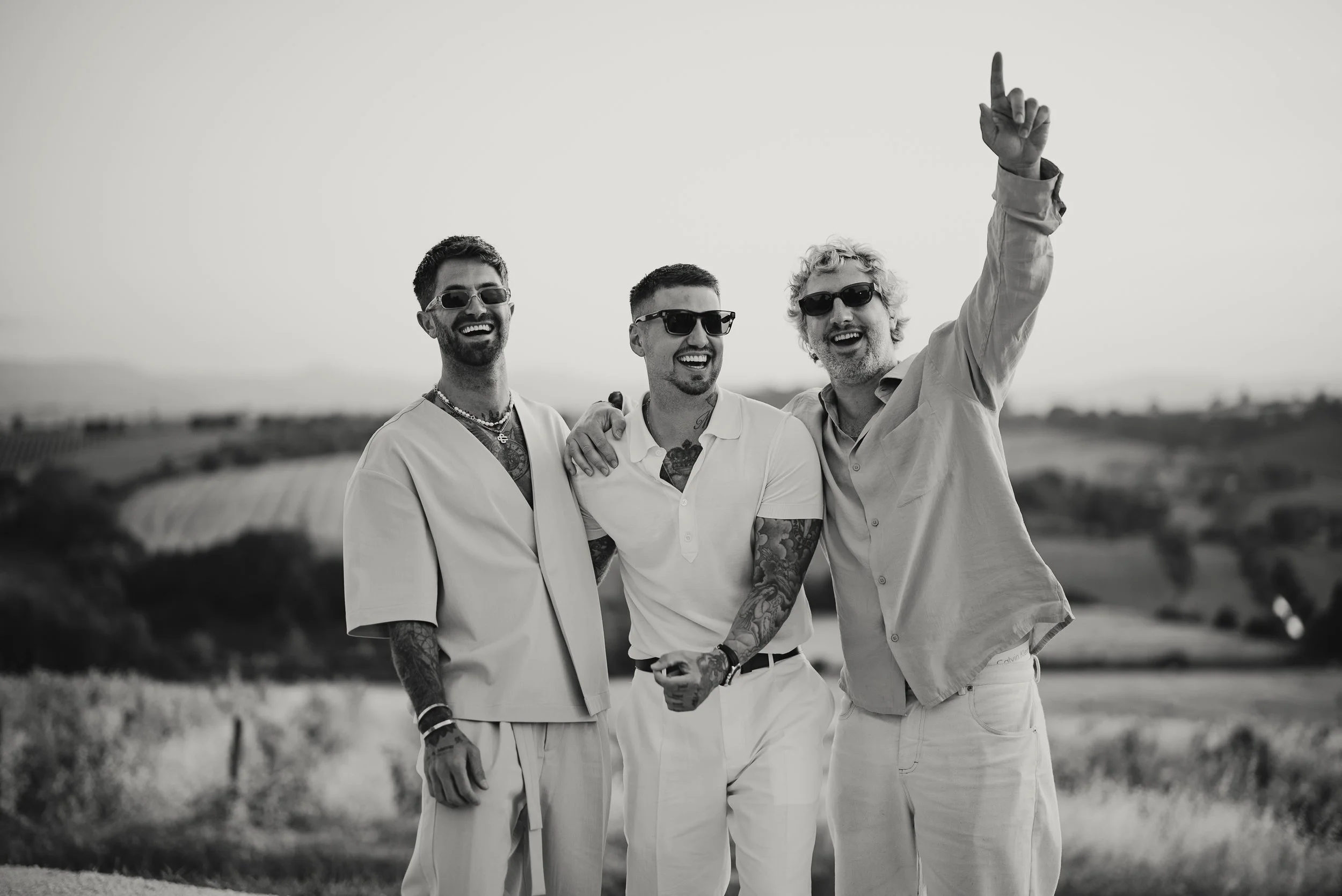 Three men wearing sunglasses and light-colored clothing smiling outdoors, with one man raising his index finger. The background is a rural landscape with fields and a distant horizon.