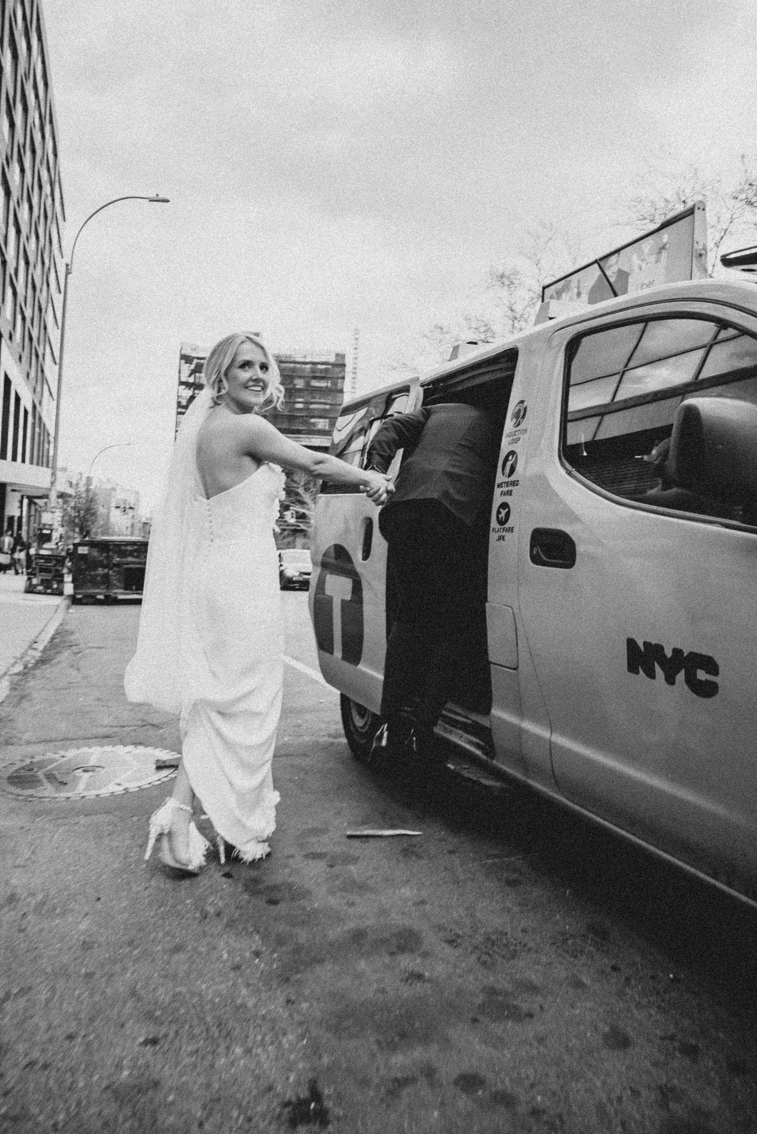 A woman in a wedding dress shaking hands with a person getting into a NYC taxi cab on a city street.
