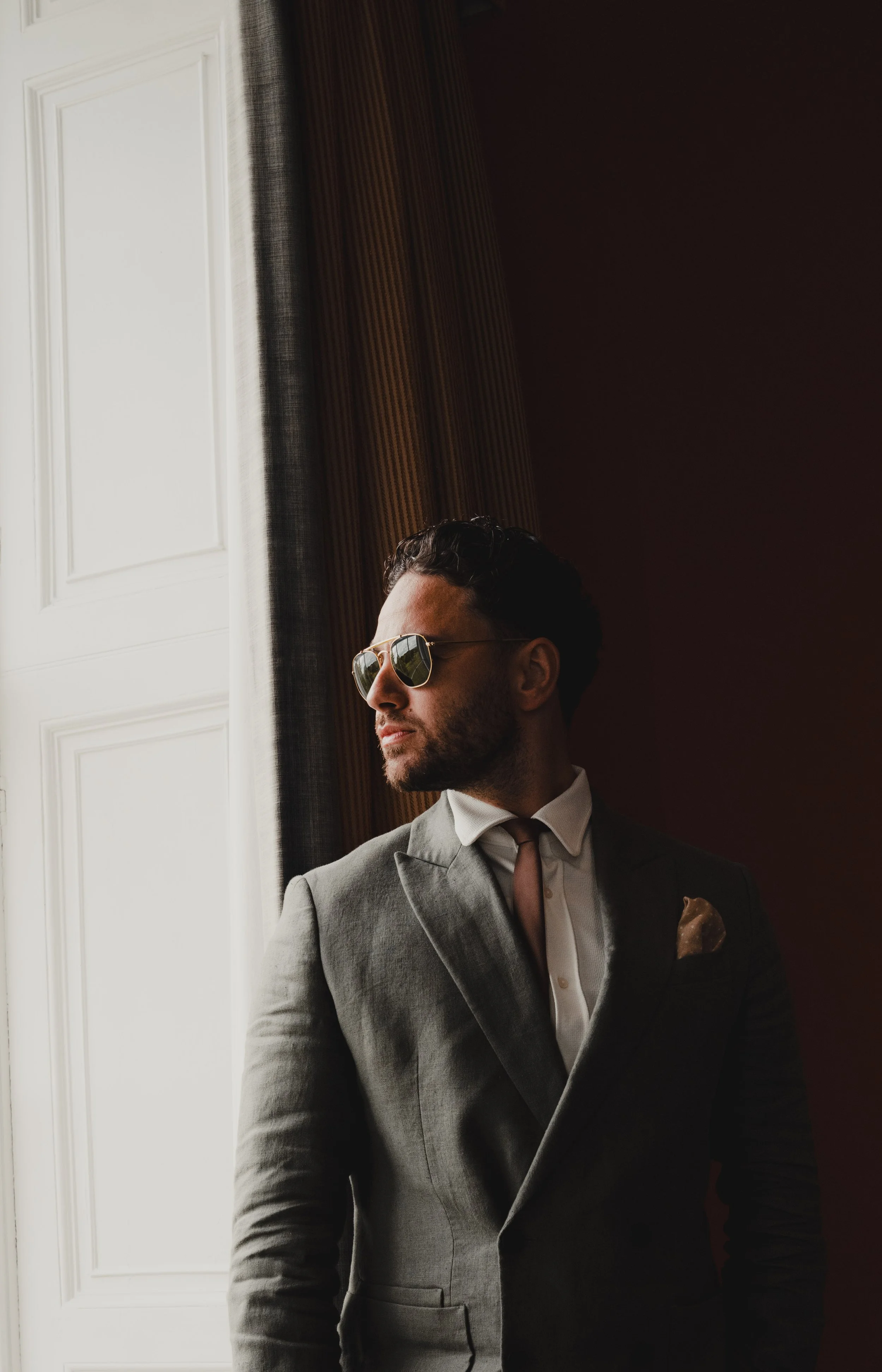 A man in a gray suit with a white shirt, brown tie, pocket square, and sunglasses, looking out a window with sunlight on his face, standing next to curtains.