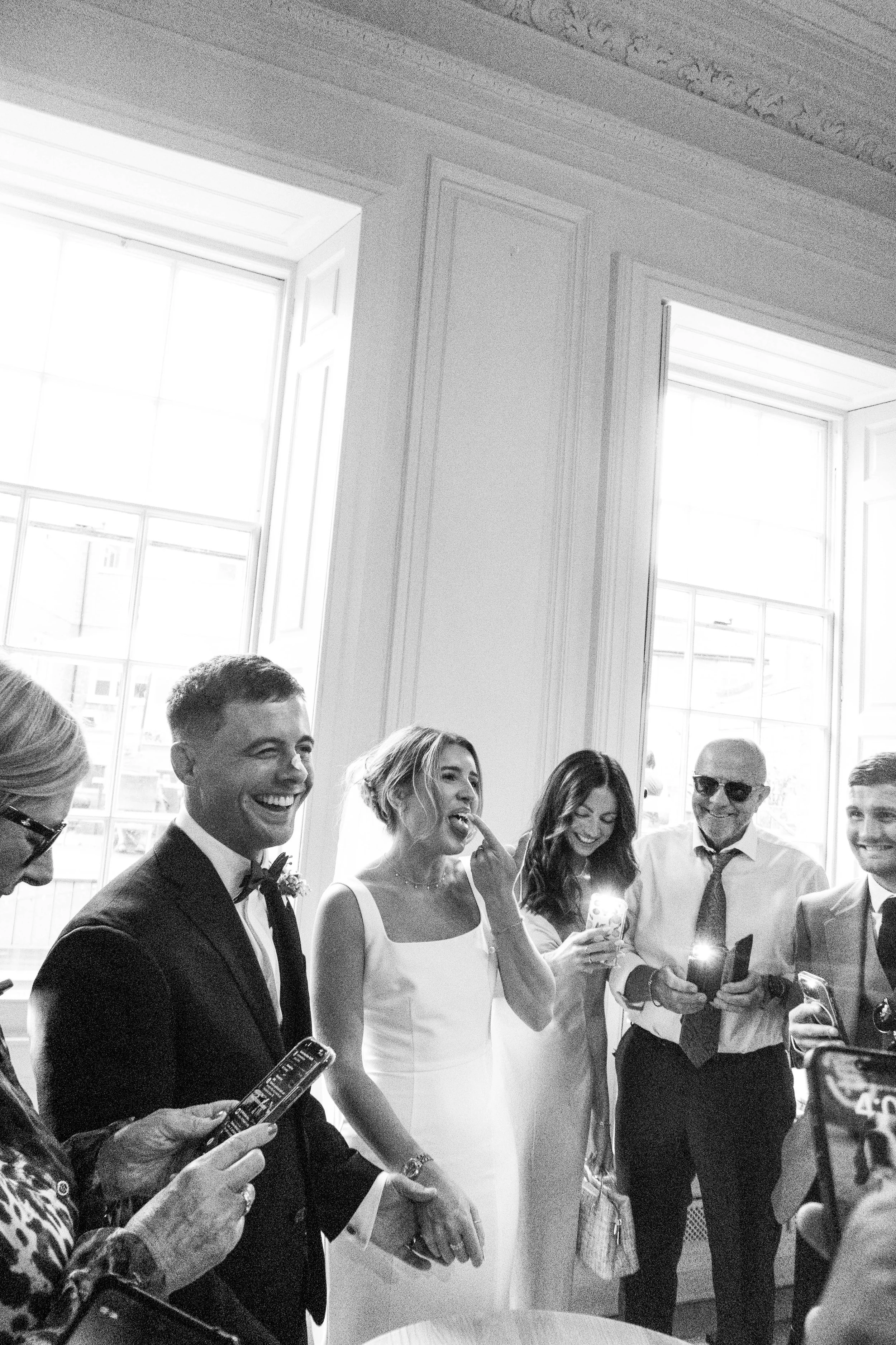 Group of people at a formal gathering, some wearing suits and a woman in a white dress, gathered indoors near large windows with sunlight, smiling and holding phones.