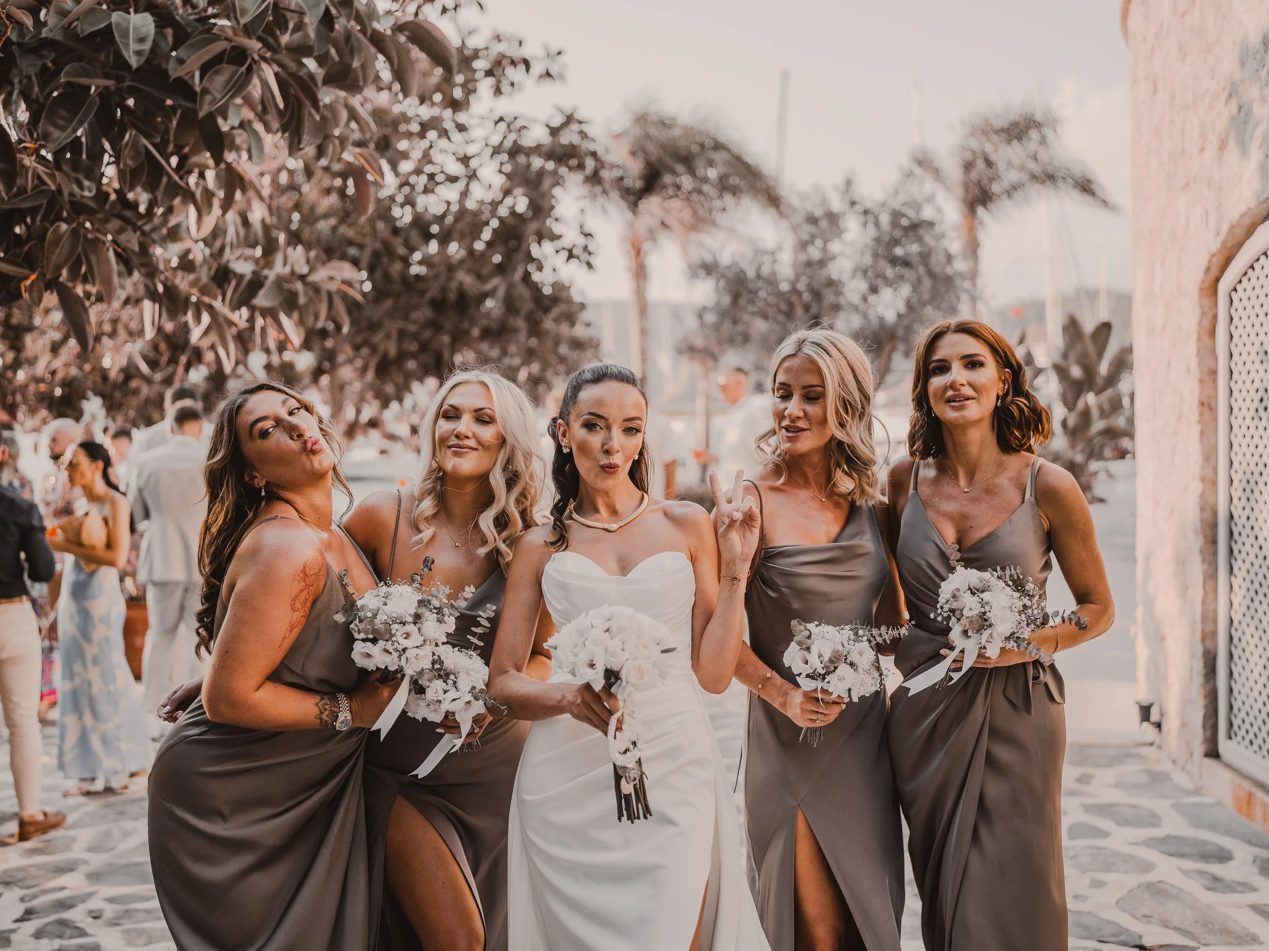 A group of women in formal dresses, with one woman in a white gown and the others in matching gray dresses, posing and making playful gestures at an outdoor event, possibly a wedding.