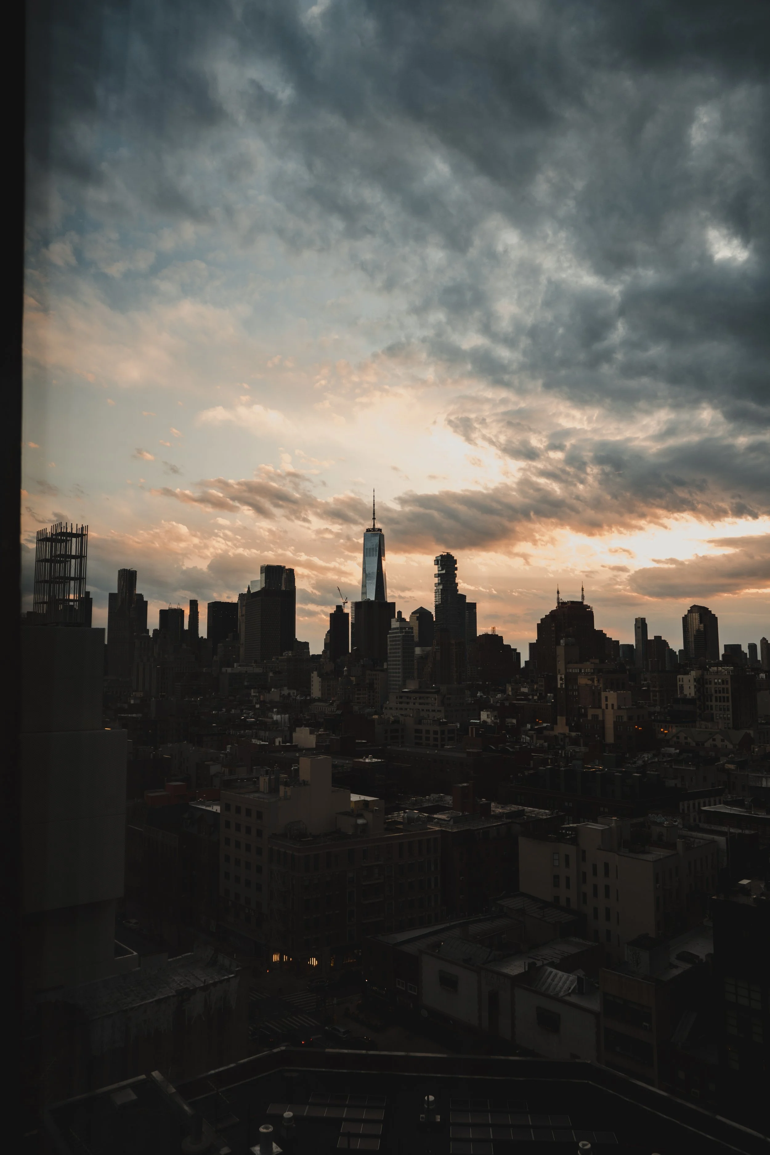 City skyline at sunset with clouds in the sky, including a prominent skyscraper with a spire, and various other tall buildings.