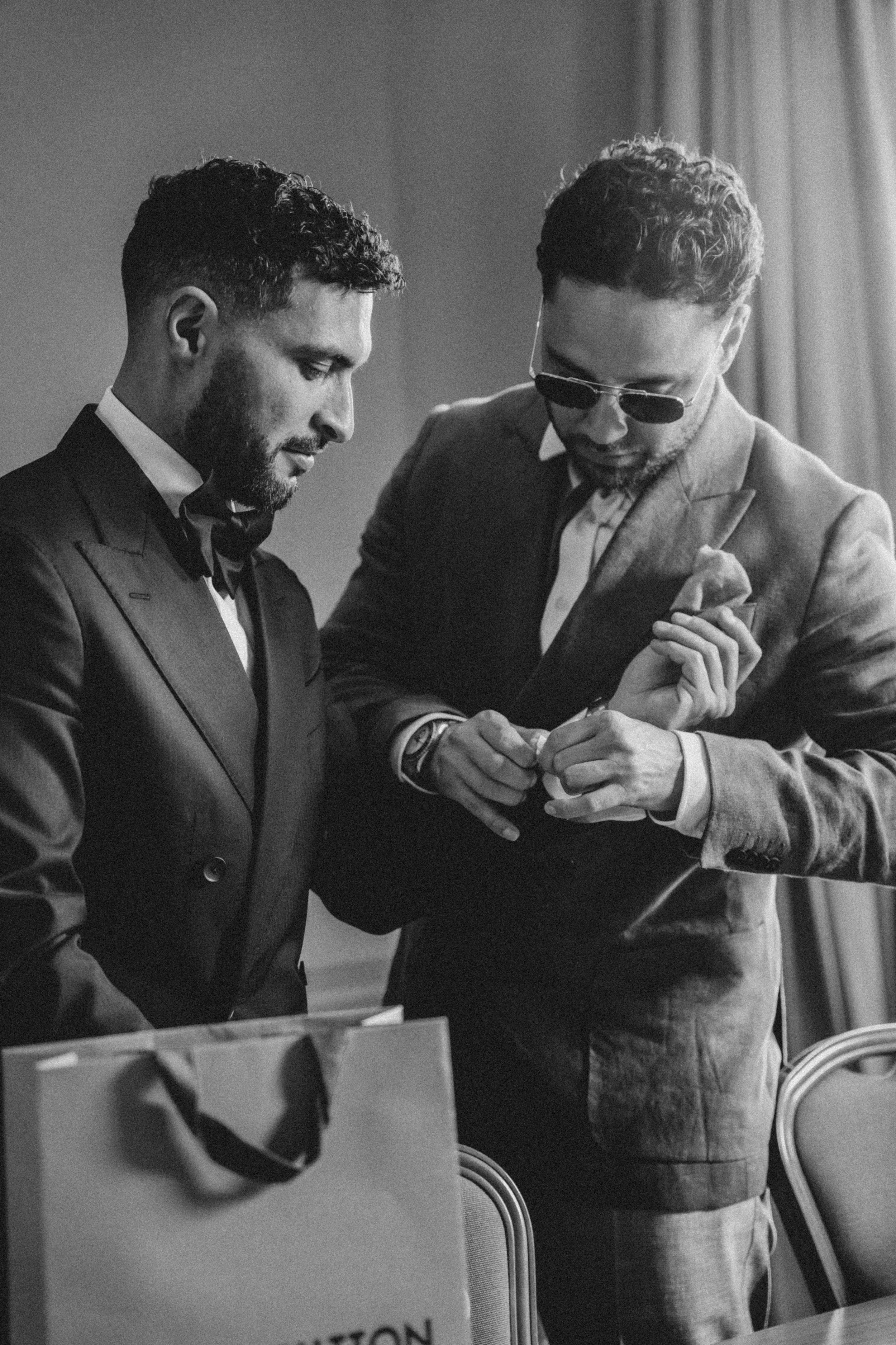 Two men in formal attire, one wearing sunglasses, interacting at a table with a box and papers, in a setting with chairs and curtains.