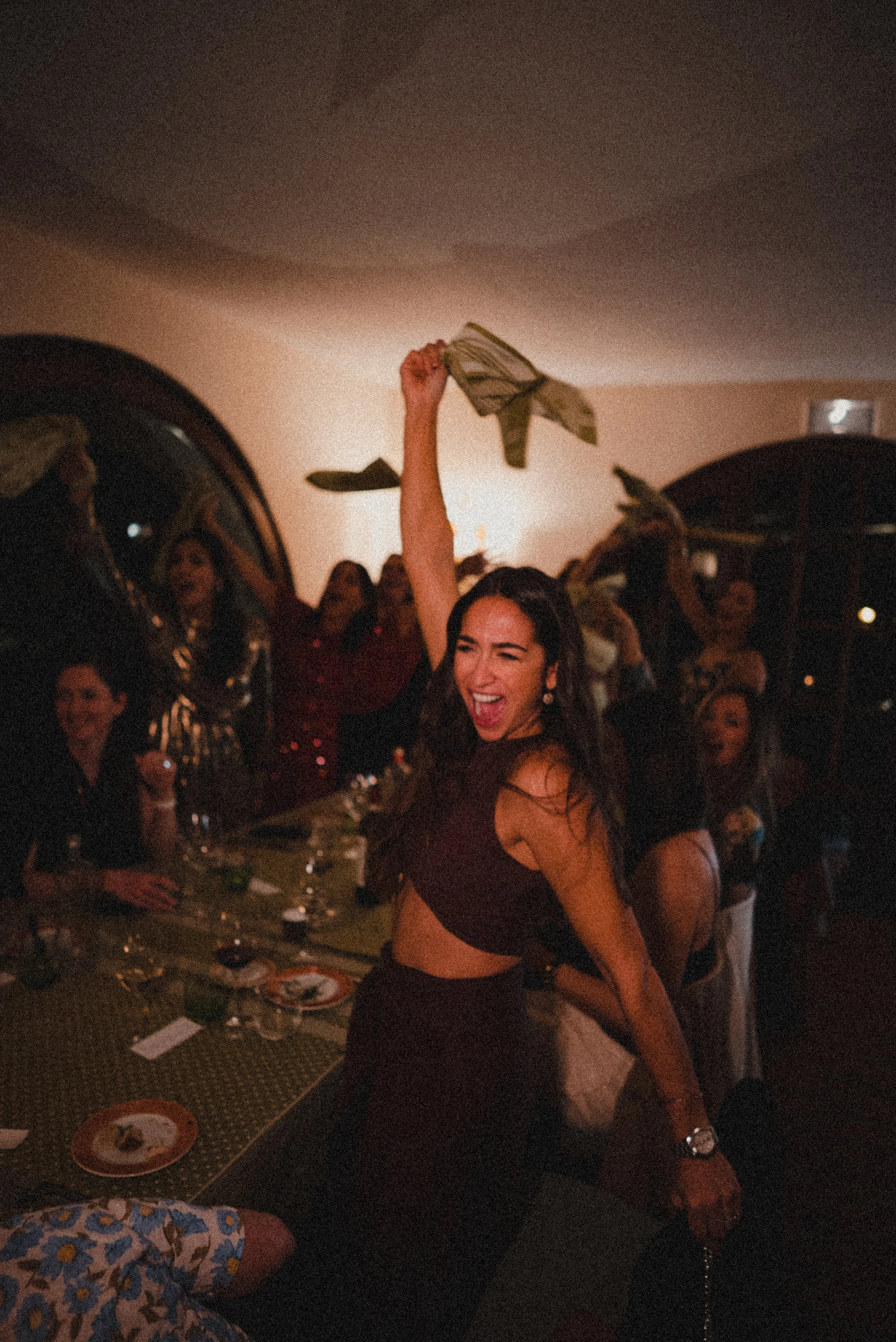 A woman is dancing and raising her arm at a lively party, surrounded by other people, with a table filled with food and drinks in the background.