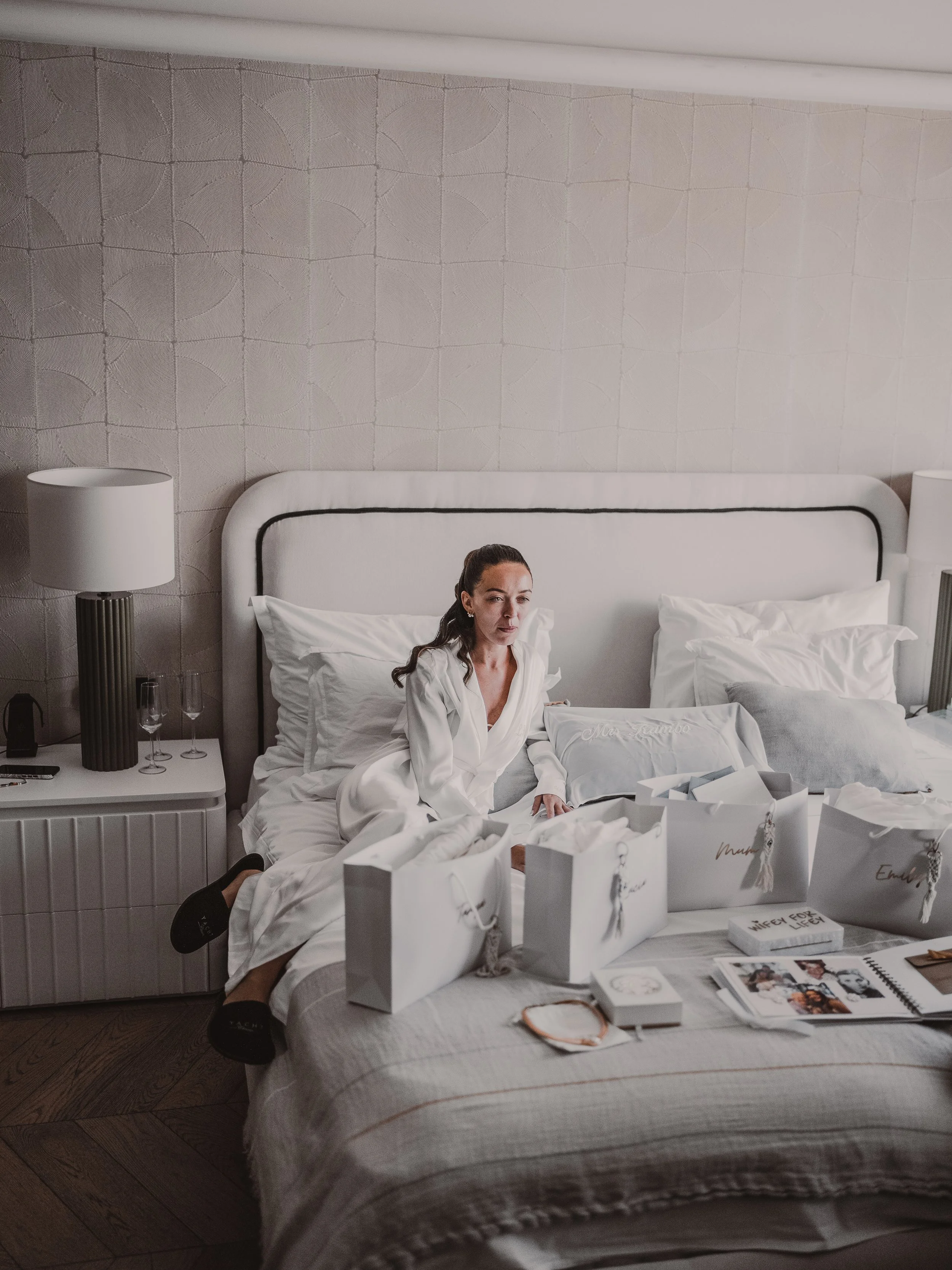 A woman with dark hair in a ponytail sitting on a bed, wearing a white robe, in a bedroom decorated in neutral tones. The bed has numerous gift bags and some books and magazines on it. There are two nightstands with lamps on either side of the bed, a