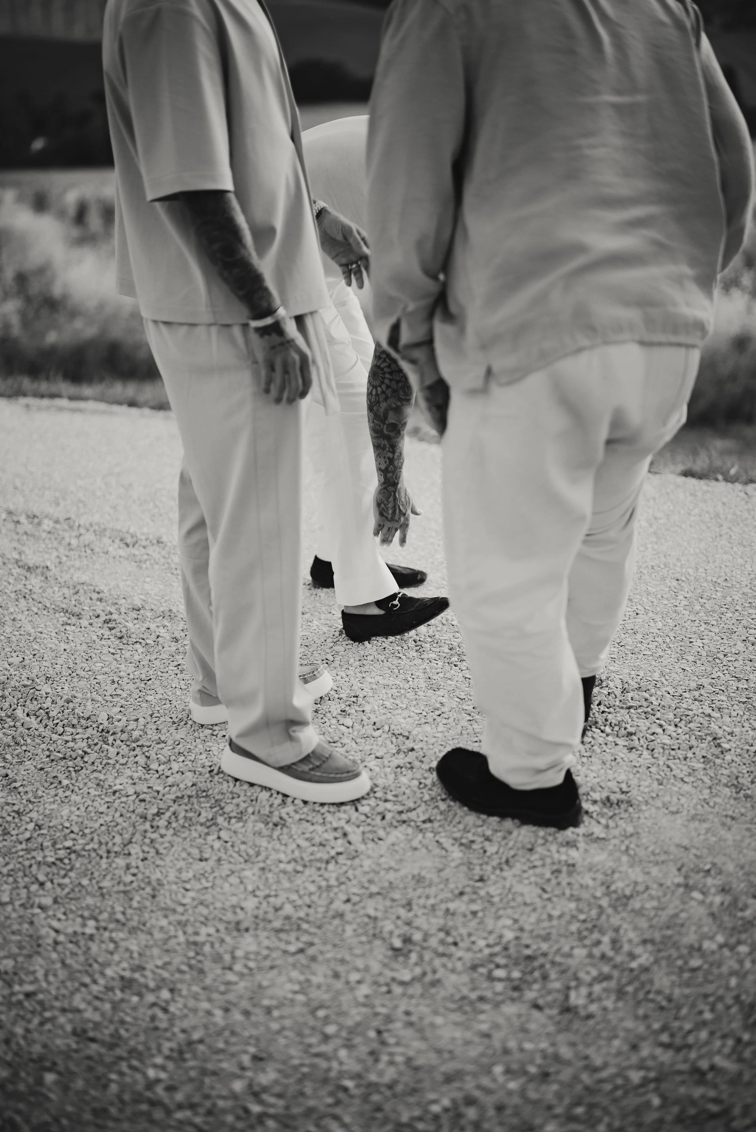 Two men are standing outdoors on a gravel surface, engaged in conversation. One man is wearing a loose, short-sleeved shirt and light-colored pants with sneakers, and has tattoos on his left arm. The second man, whose back is to the camera, is dresse