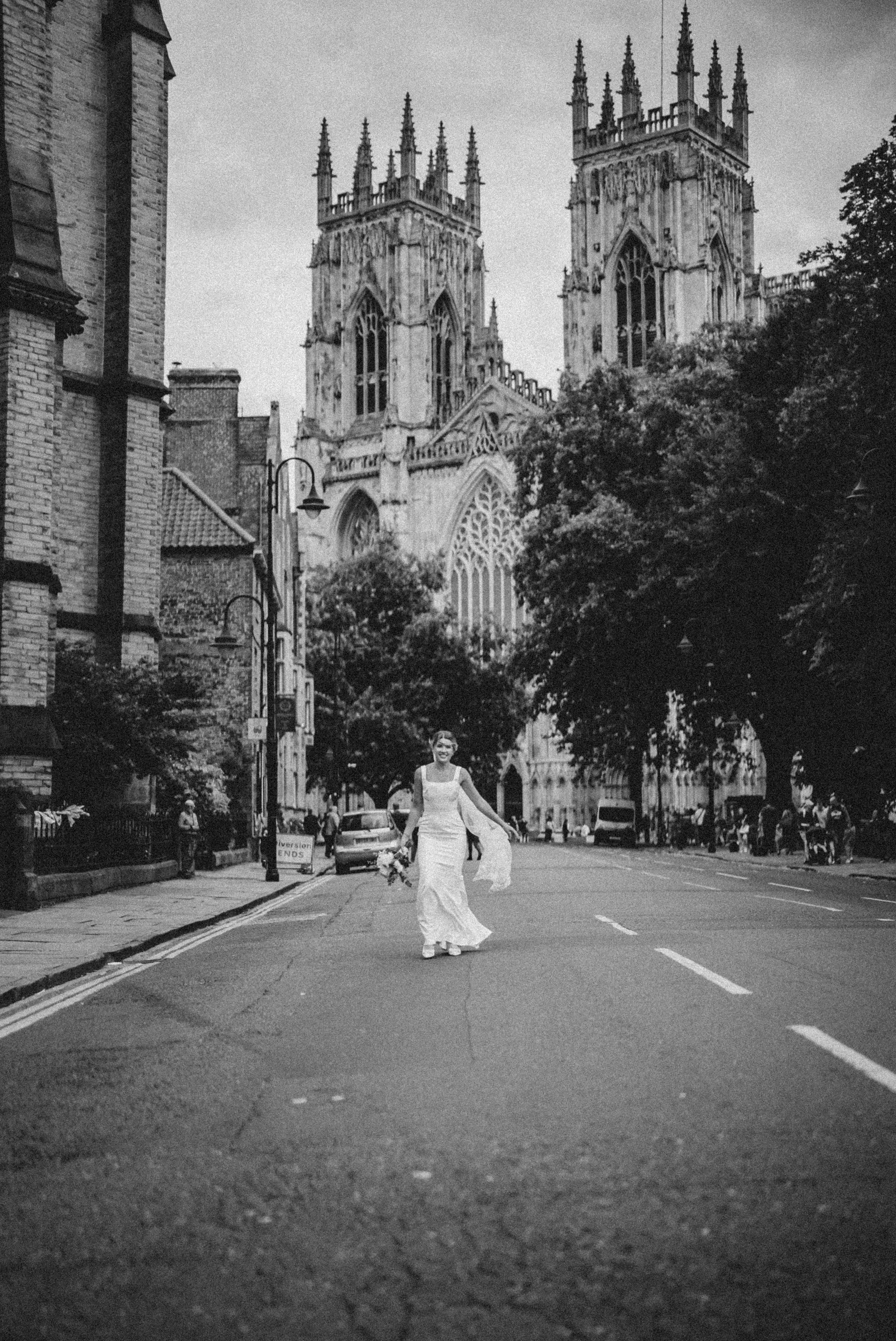 A woman in a wedding dress walking down a city street holding a bouquet of flowers with tall historic church towers in the background.