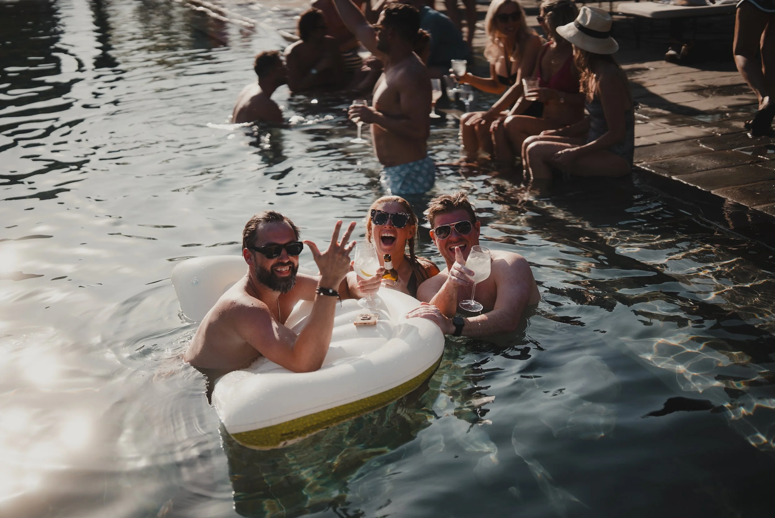 A group of people enjoying a pool party, sitting and floating in the water, smiling, wearing sunglasses, and holding drinks during the daytime.