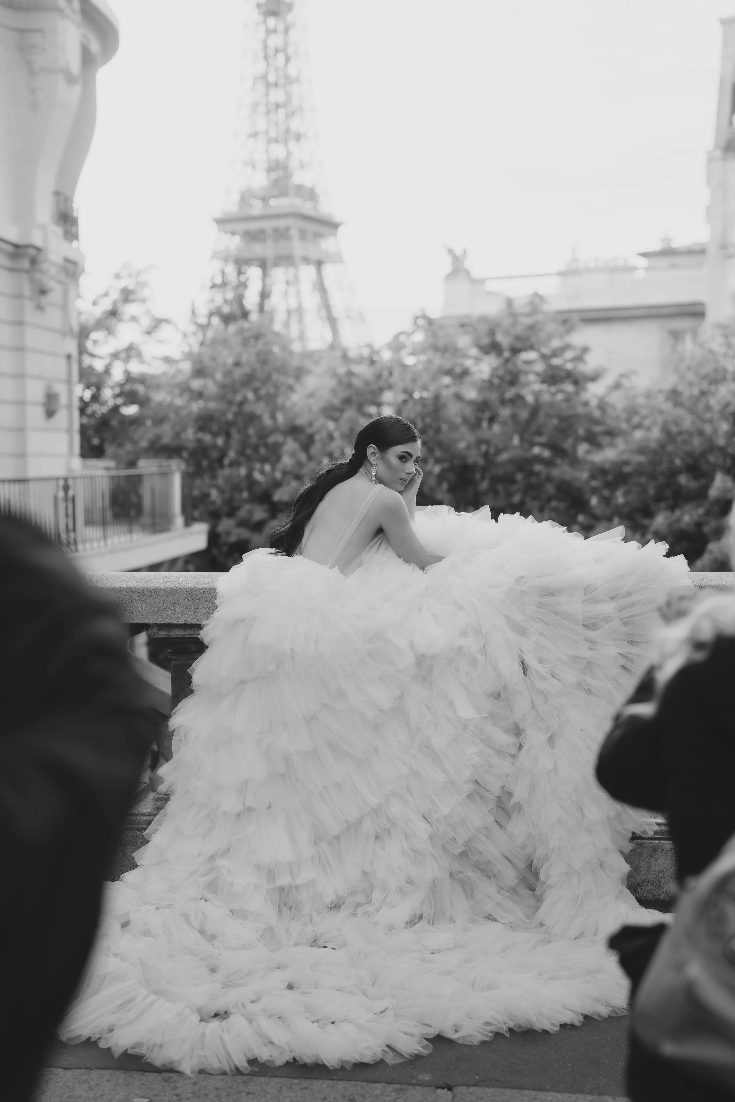 A woman in a dramatic white gown with a large ruffled skirt, seated outdoors with a cityscape and a tower in the background.