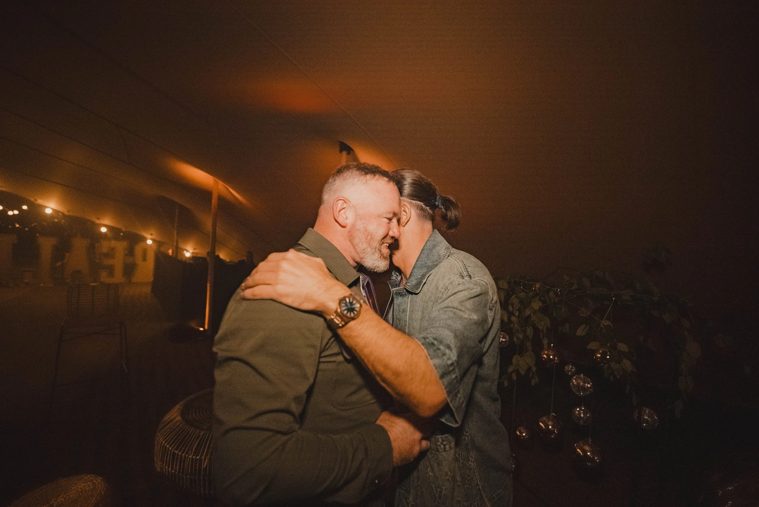 Two men are hugging and smiling at each other inside a dimly lit space with warm lighting. One man has a beard and short hair, and the other has long hair tied back. There is a basketball hoop and some decorations in the background.