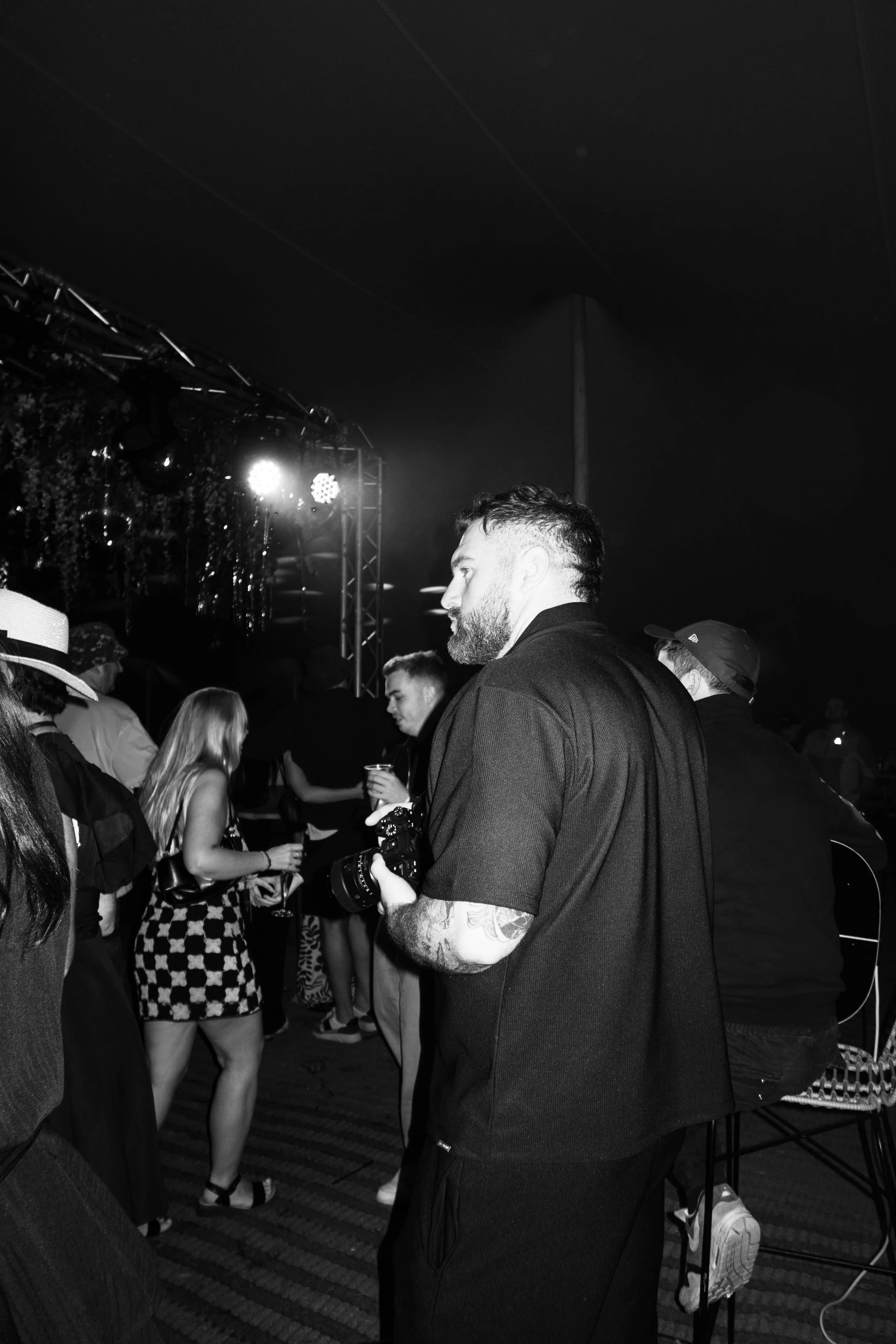 A man with a beard and tattoos on his arm holding a camera in a crowded indoor event with people mingling and stage lighting in the background.