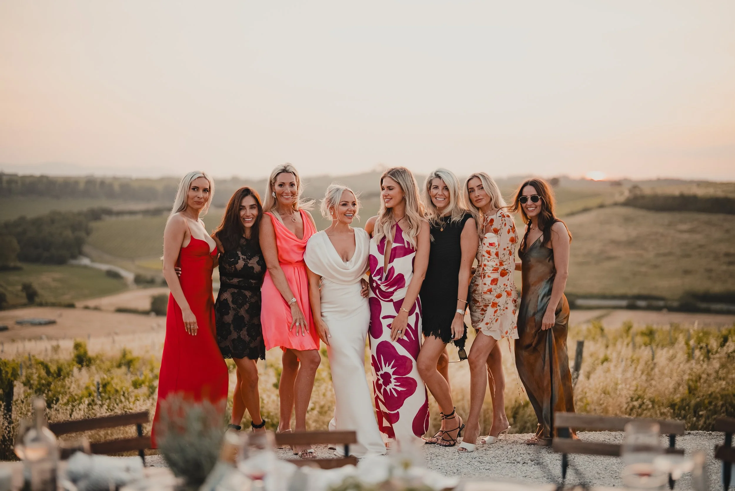 A group of nine women in colorful dresses standing outdoors in a vineyard at sunset, smiling and posing for a photograph.