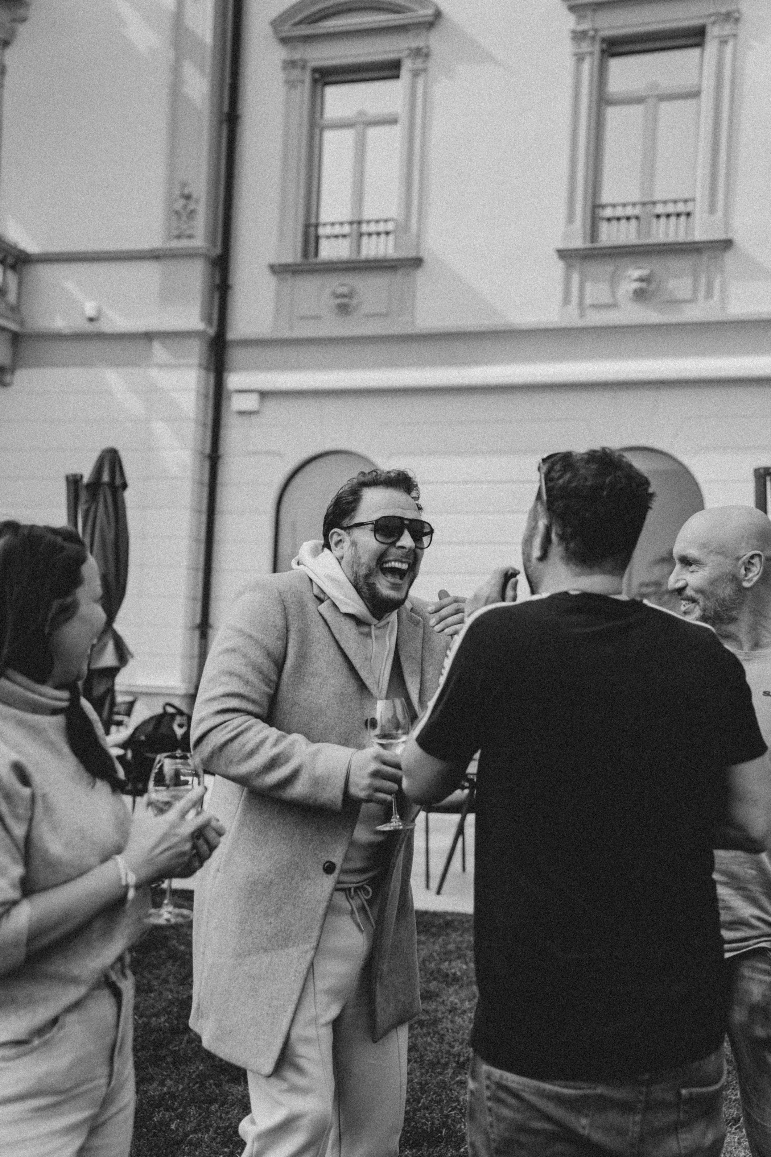 A group of people at an outdoor social gathering, engaged in conversation, holding wine glasses, with a building featuring windows in the background.
