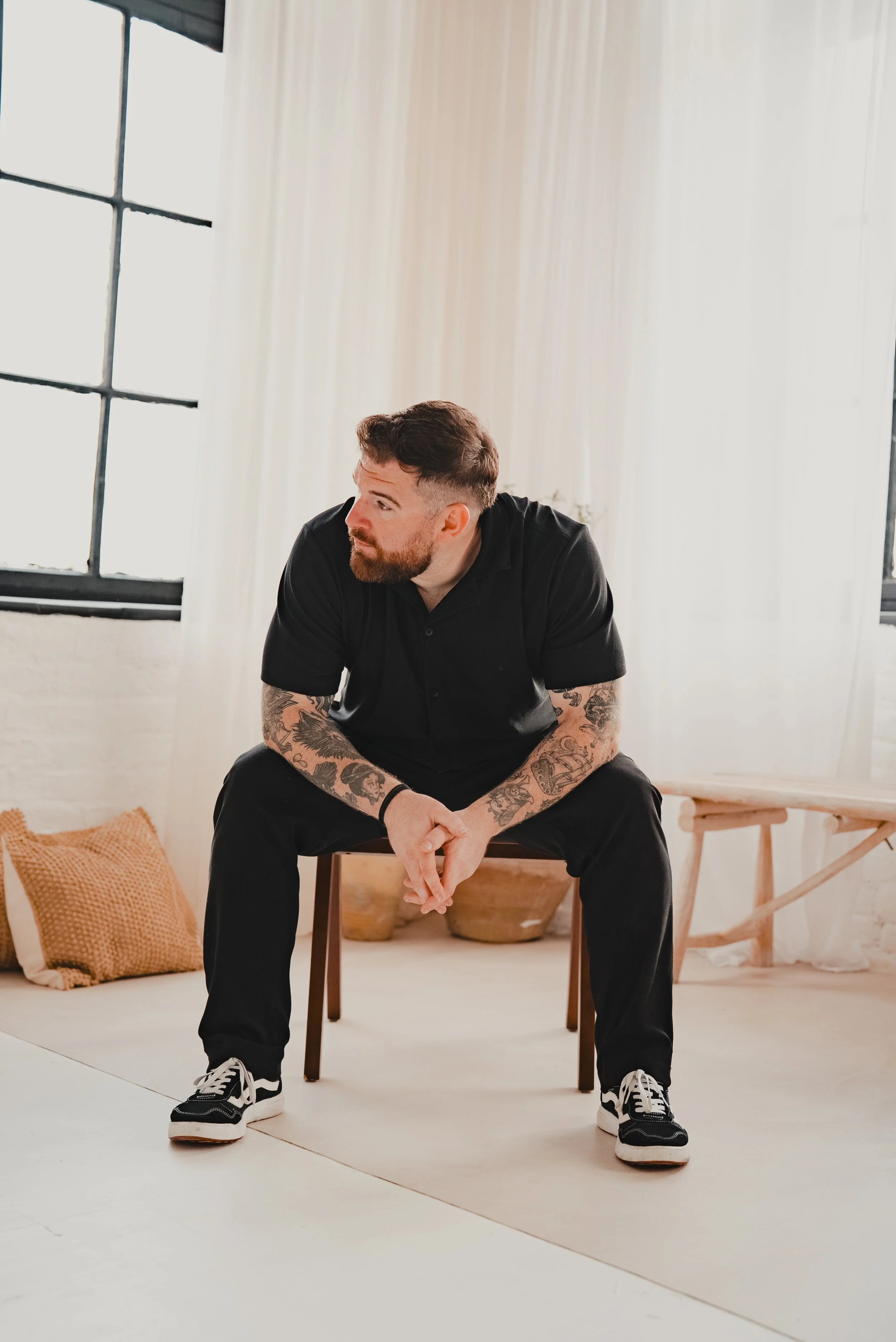 A man with tattoos on his arms, beard, and styled hair, sitting on a wooden chair in a bright room with large windows, white curtains, a beige pillow on the floor, and decorative clay pots.
