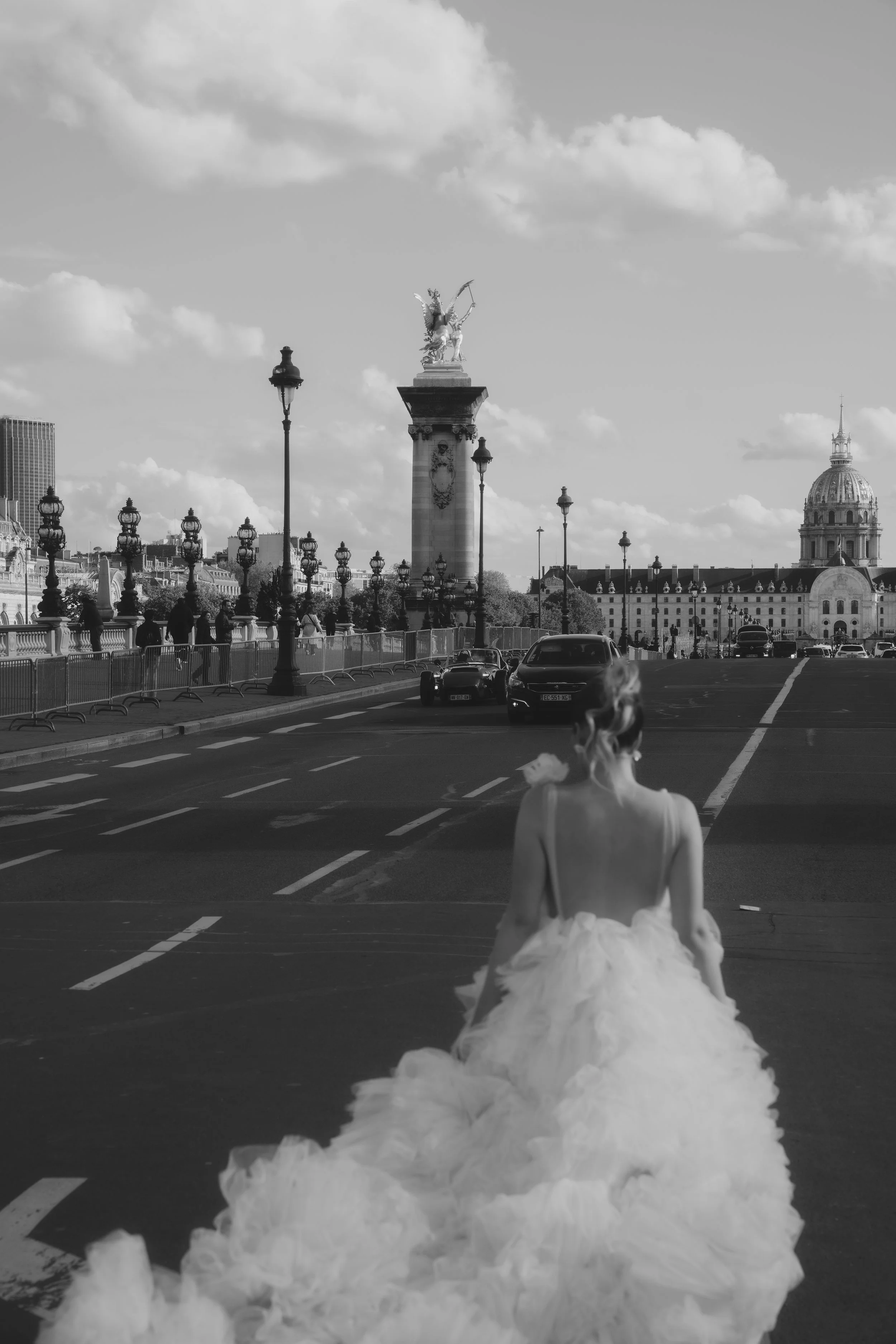 A woman in a white dress sitting on a street with a large, billowy skirt, facing away from the camera. In the background, there is a cityscape with tall buildings, lamp posts, a monument with an angel on top, and a dome building. The scene is in blac