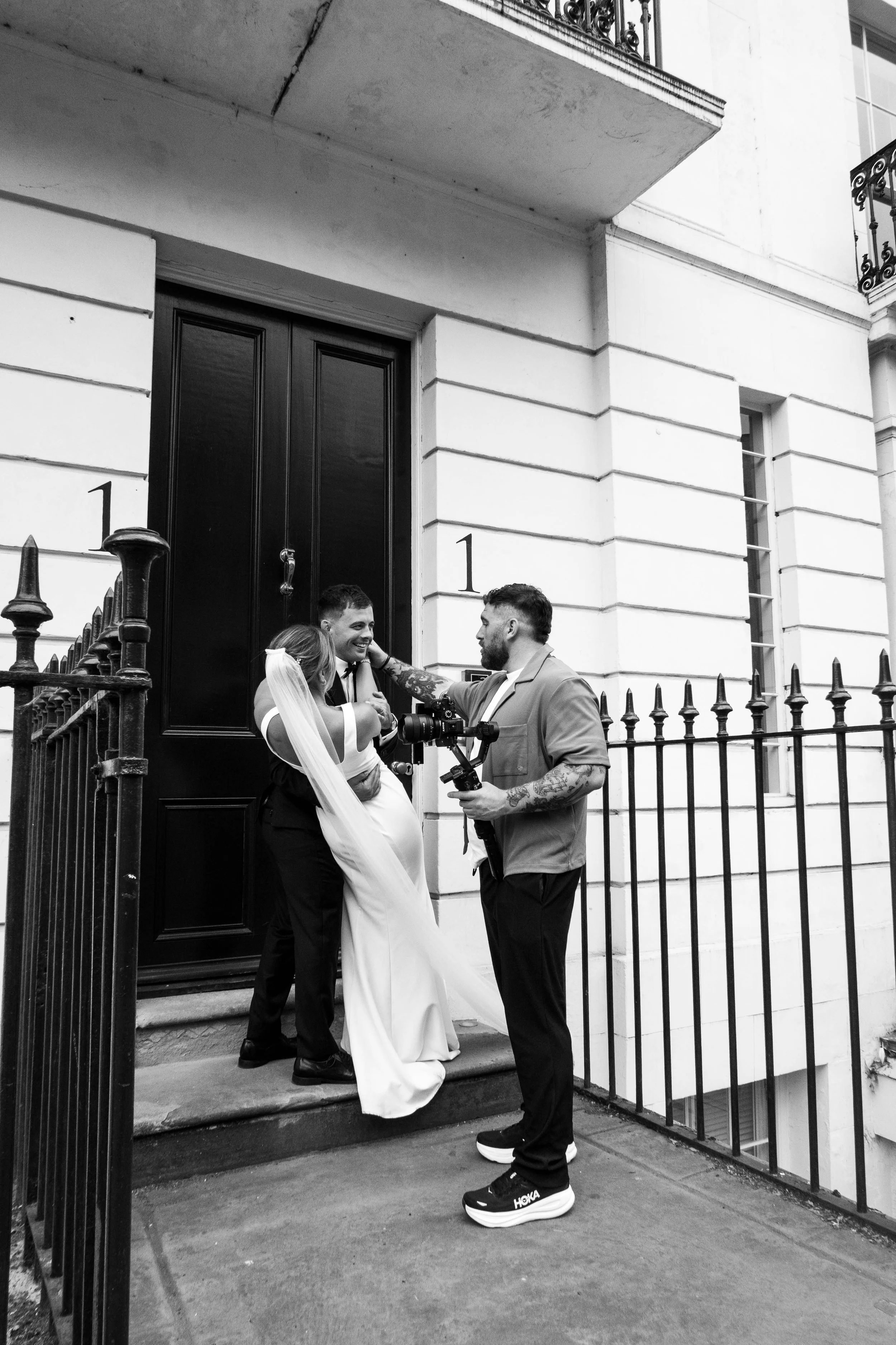 A wedding ceremony taking place outside a building with a black door. A bride in a white dress and veil is hugging a groom who is dressed in black. A photographer is photographing the couple, wearing a light-colored shirt and black sneakers with the word 'HOKA' on the sole. The scene is behind a black iron fence.