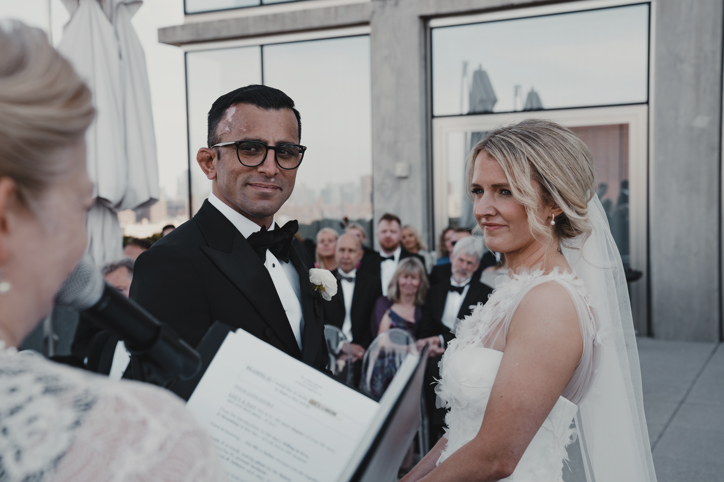 A wedding ceremony on a rooftop with a man in a tuxedo and a woman in a white wedding dress standing close together, surrounded by guests, with a person speaking into a microphone in the foreground.