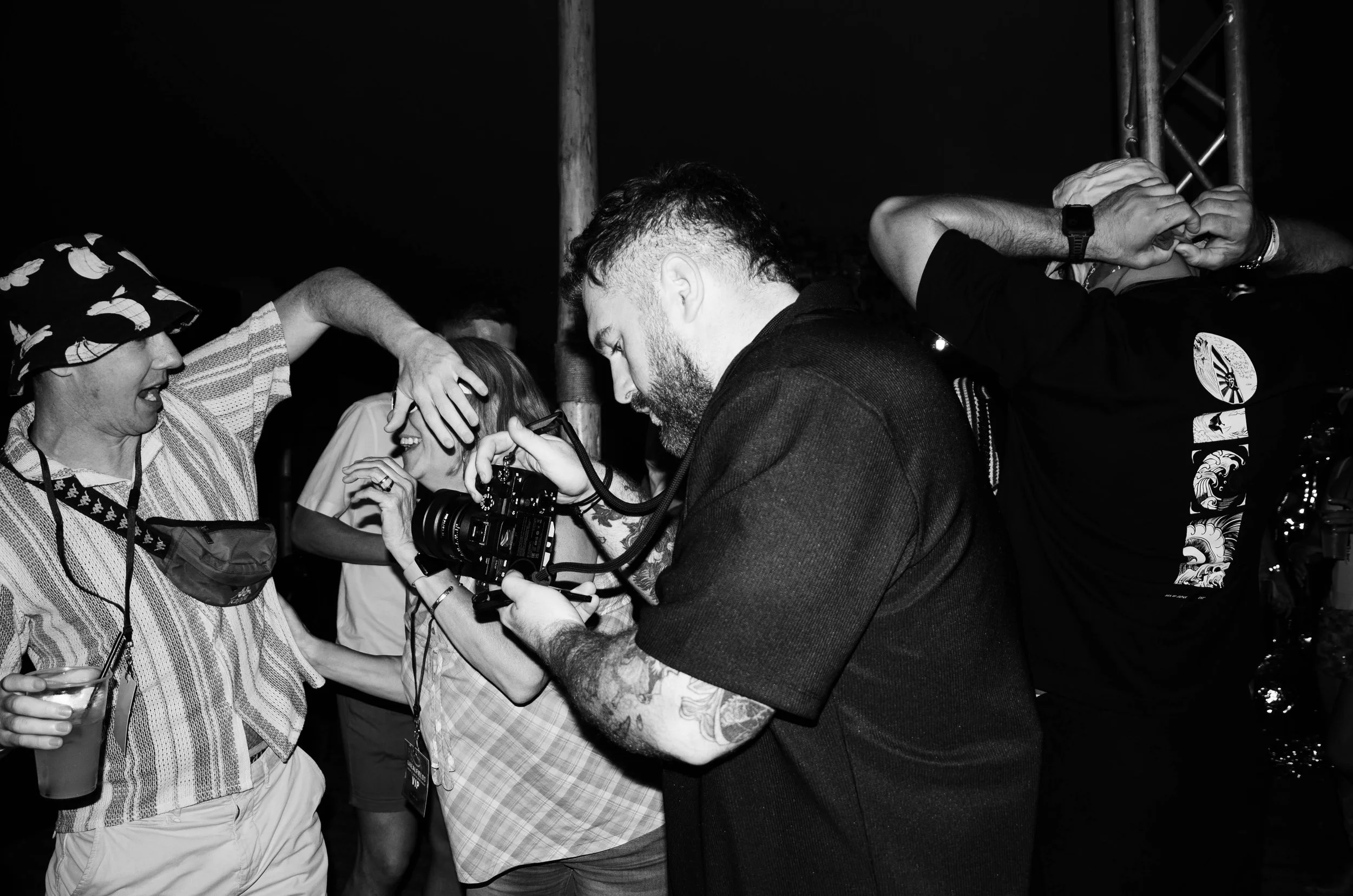A group of people at a social event, with three individuals in focus, engaged in conversation and taking photos. One person is wearing a patterned hat, another has a beard and is holding a camera, and the third is also taking a photo with their phone