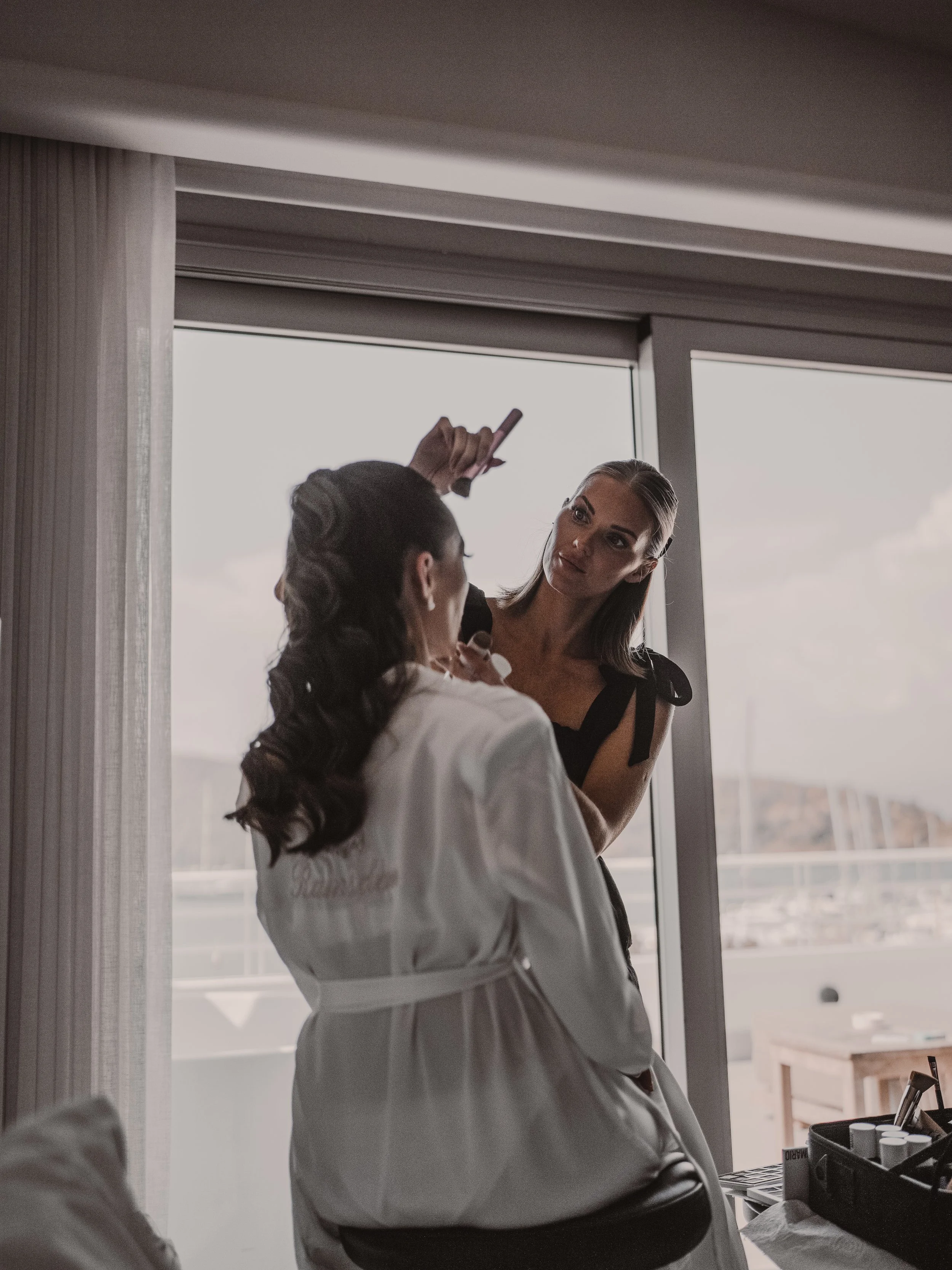 A woman applying makeup on another woman, who is sitting near a large window with a city view.