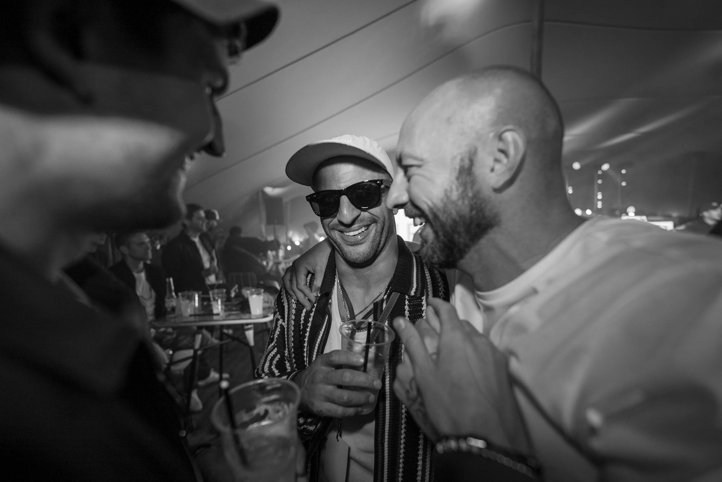 Four men are smiling and laughing while holding drinks at a social gathering in a dimly lit indoor setting.