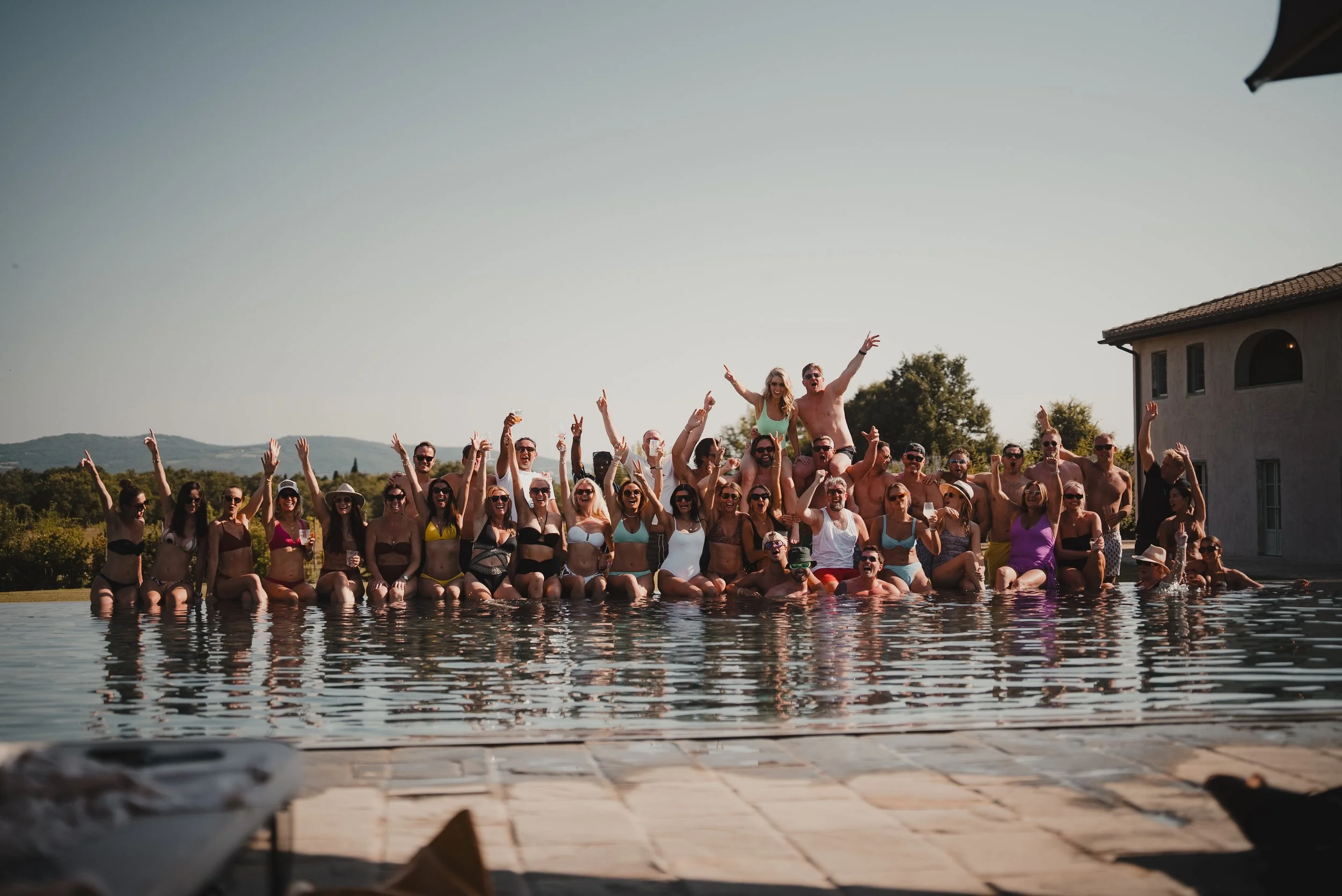 A large group of people in swimsuits gathering at the edge of a swimming pool, celebrating and waving, with some people sitting on others' shoulders, sunny weather, and a house and trees in the background.