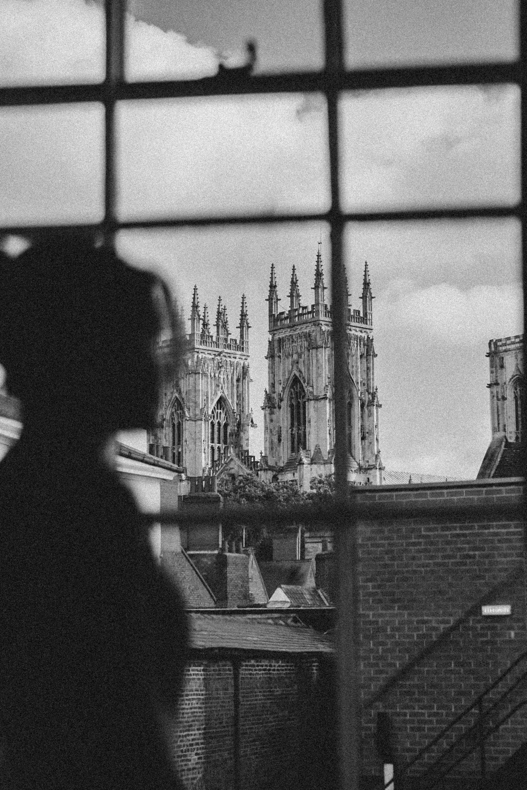 Black and white photo showing a church with Gothic architecture, viewed through a window with a grid pattern. In the foreground, there are darker out-of-focus shapes, possibly city rooftops and brick buildings.