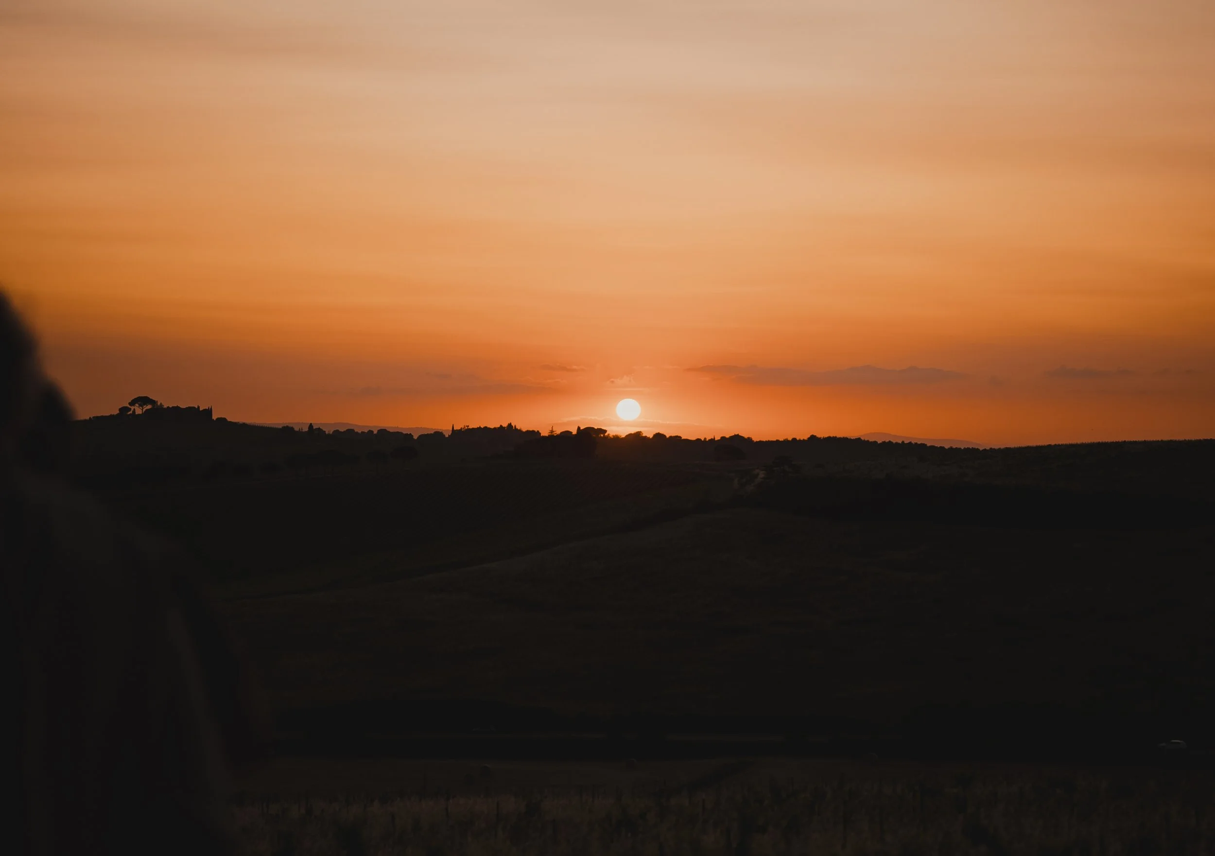 Sun setting on a rural landscape with trees and hills, casting a warm orange glow across the sky.