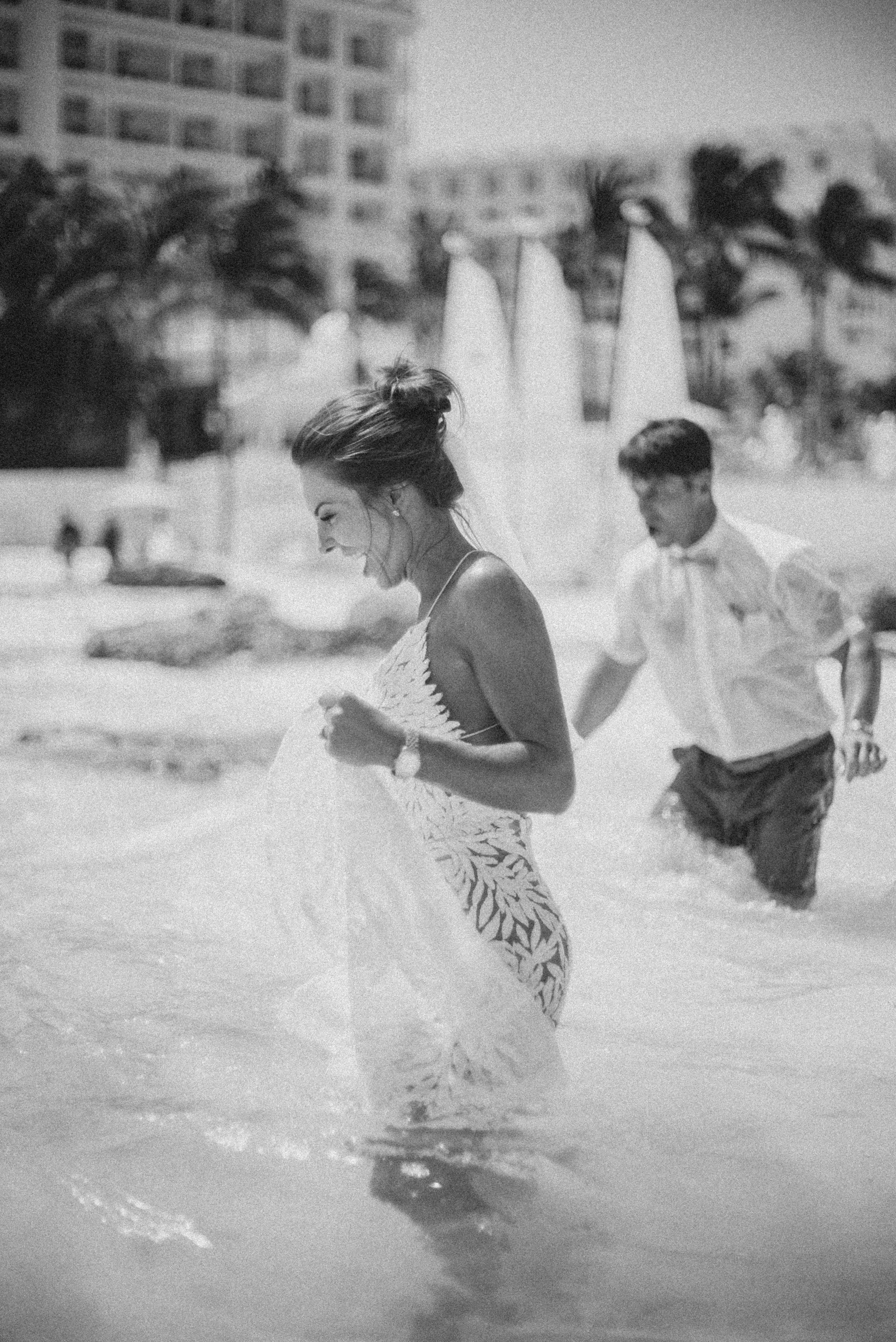 A young woman with dark hair styled in a loose updo wearing a patterned dress is walking in a shallow pool of water on a sunny day. She is smiling and looking down. Behind her, a young man with dark hair and glasses, dressed in a button-up shirt and shorts, is walking in the same direction. In the background, there are water fountains, palm trees, and a multi-story building.